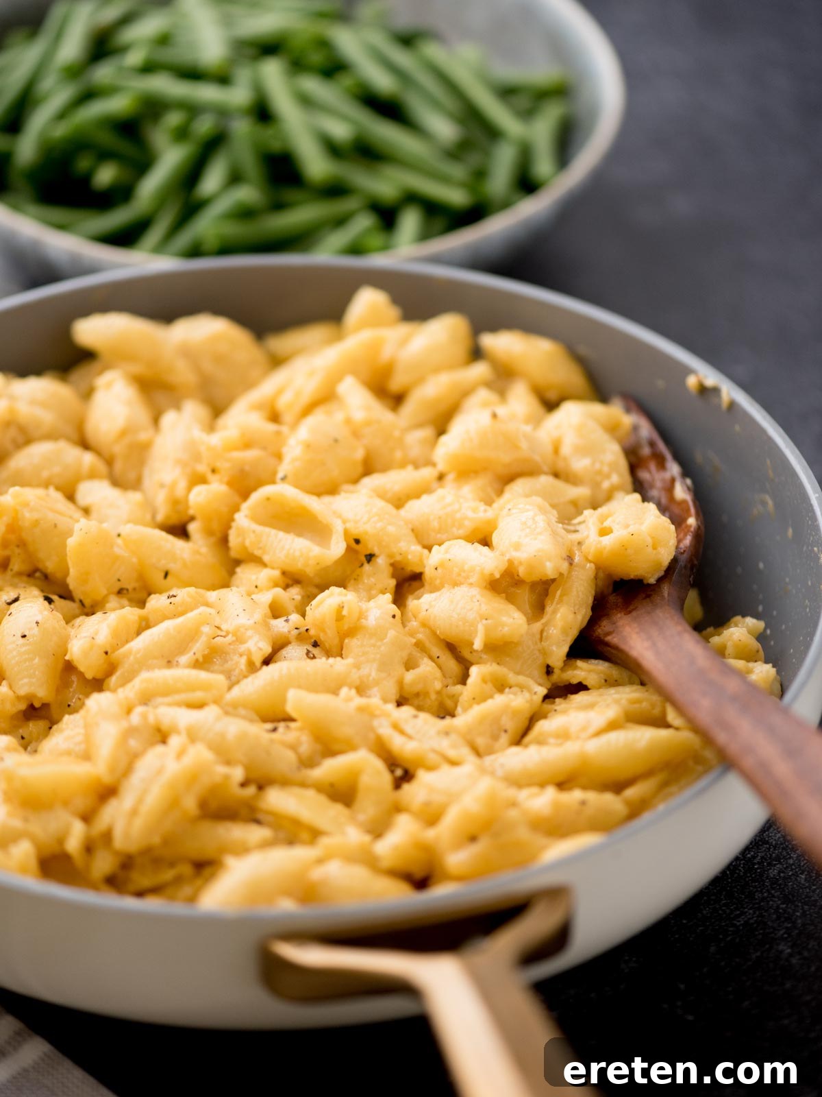 A skillet filled with creamy macaroni and cheese, with a wooden spoon resting inside. In the background, a bowl of fresh green beans is partially visible. Both dishes are placed on a dark surface, creating an inviting comfort food scene.