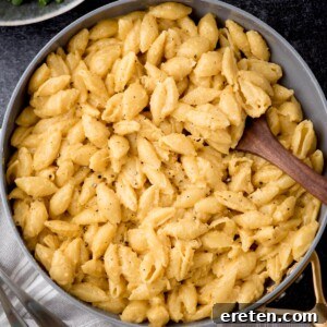 A skillet filled with creamy mac and cheese pasta shells, garnished with cracked black pepper. A wooden spoon rests in the dish, ready to serve. The background shows a hint of a green leafy side dish.