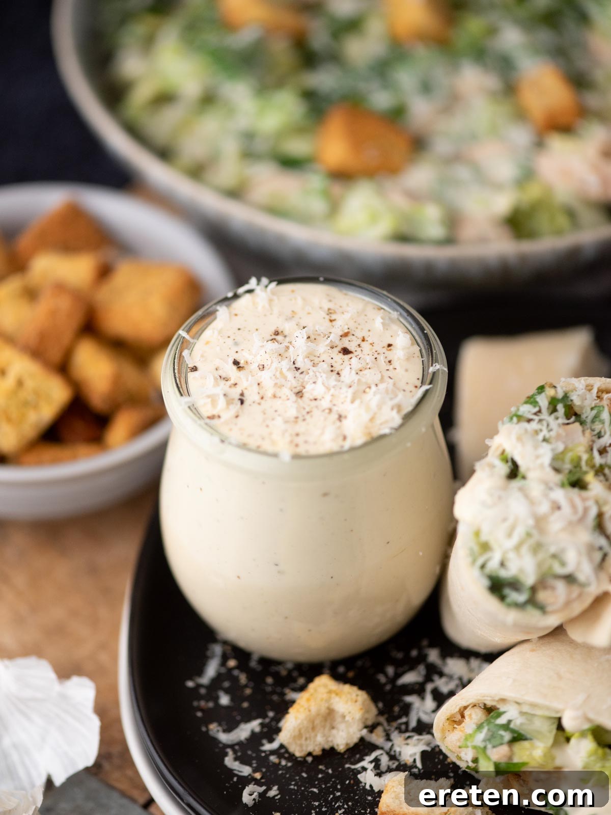 A jar of creamy Caesar salad dressing is surrounded by croutons, a wrap filled with salad, and a bowl of Caesar salad in the background. Grated cheese is sprinkled on top of the dressing and the wrap.