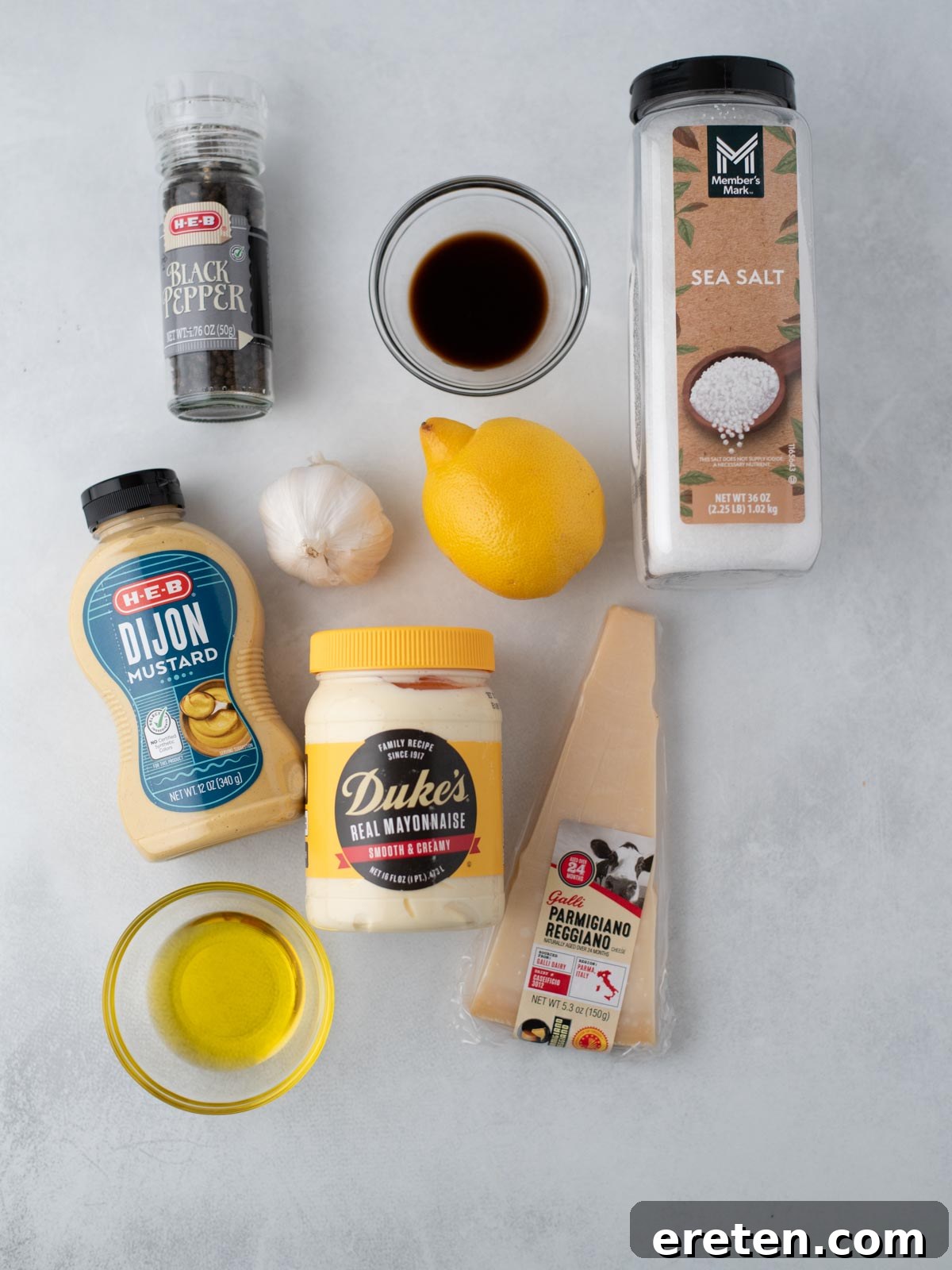 Ingredients for Caesar dressing displayed on a light countertop, including black pepper, Worcestershire sauce, sea salt, Dijon mustard, mayonnaise, a lemon, garlic bulbs, olive oil, and a wedge of Parmesan cheese.