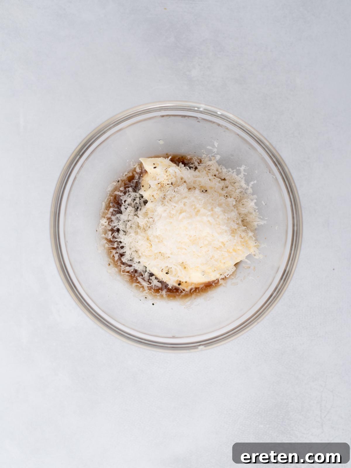 A glass bowl containing grated cheese, mayonnaise, Worcestershire sauce, and black pepper, set on a light gray surface.