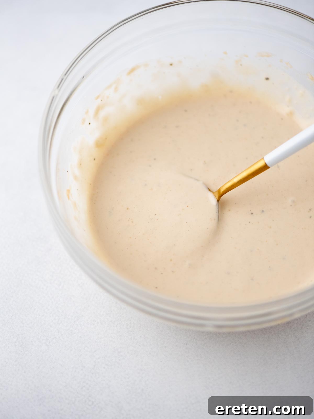 A glass bowl filled with creamy, light beige sauce or dressing, being mixed with a white and gold spoon on a light surface.