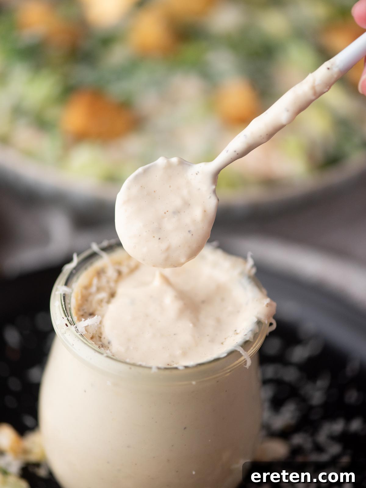 A close-up of a jar filled with creamy Caesar salad dressing, with a spoon lifting some of the dressing above the jar. The background shows a blurred bowl of salad.