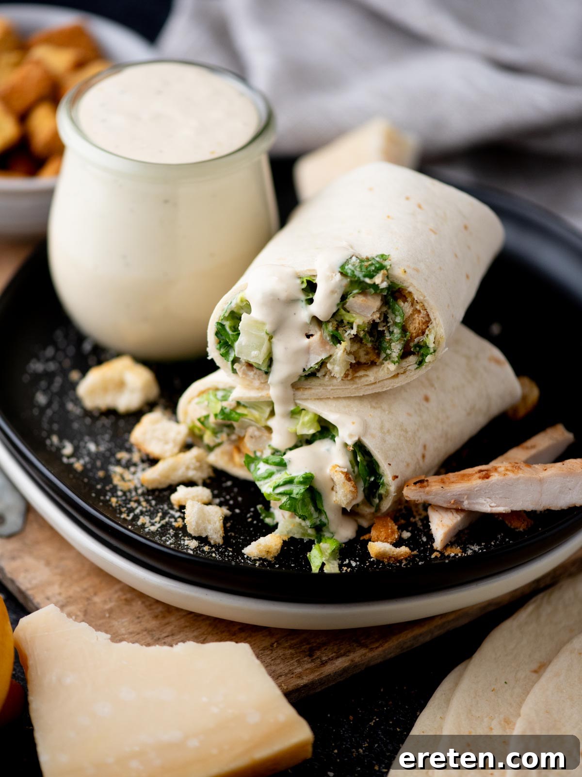 A Caesar salad wrap cut in half sits on a black plate, filled with lettuce, chicken, croutons, and creamy Caesar dressing, with extra dressing in a jar nearby and sprinkled cheese on the plate.