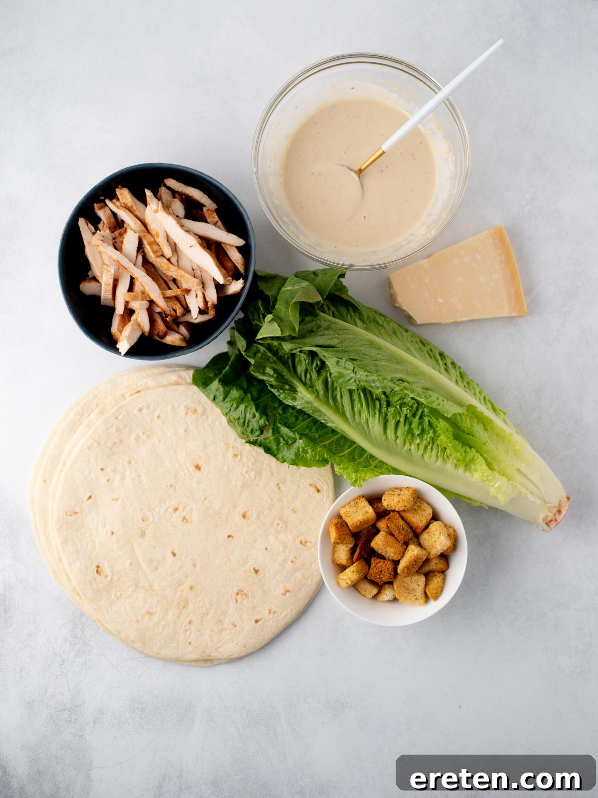 Ingredients for a chicken Caesar wrap: flour tortillas, sliced grilled chicken, romaine lettuce, Caesar dressing in a bowl, Parmesan cheese wedge, and a small bowl of croutons on a white surface.