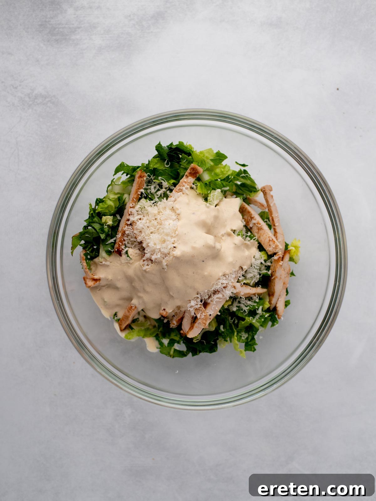 A glass bowl filled with chopped romaine lettuce, grilled chicken strips, grated cheese, and a generous amount of creamy dressing on top, set against a light gray background.