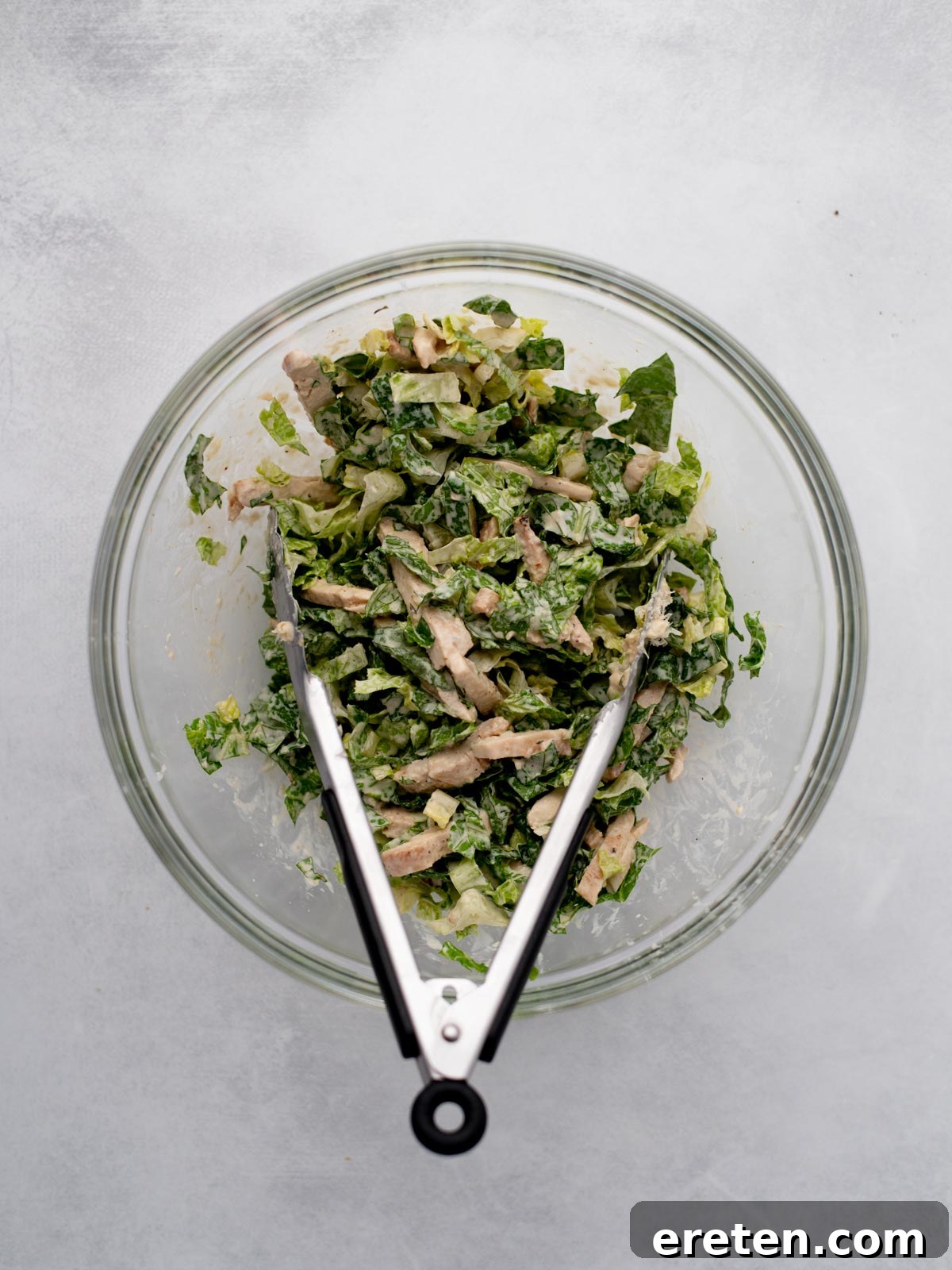 A glass bowl filled with chopped salad greens and pieces of cooked chicken, with metal tongs resting inside the bowl, on a light gray surface.