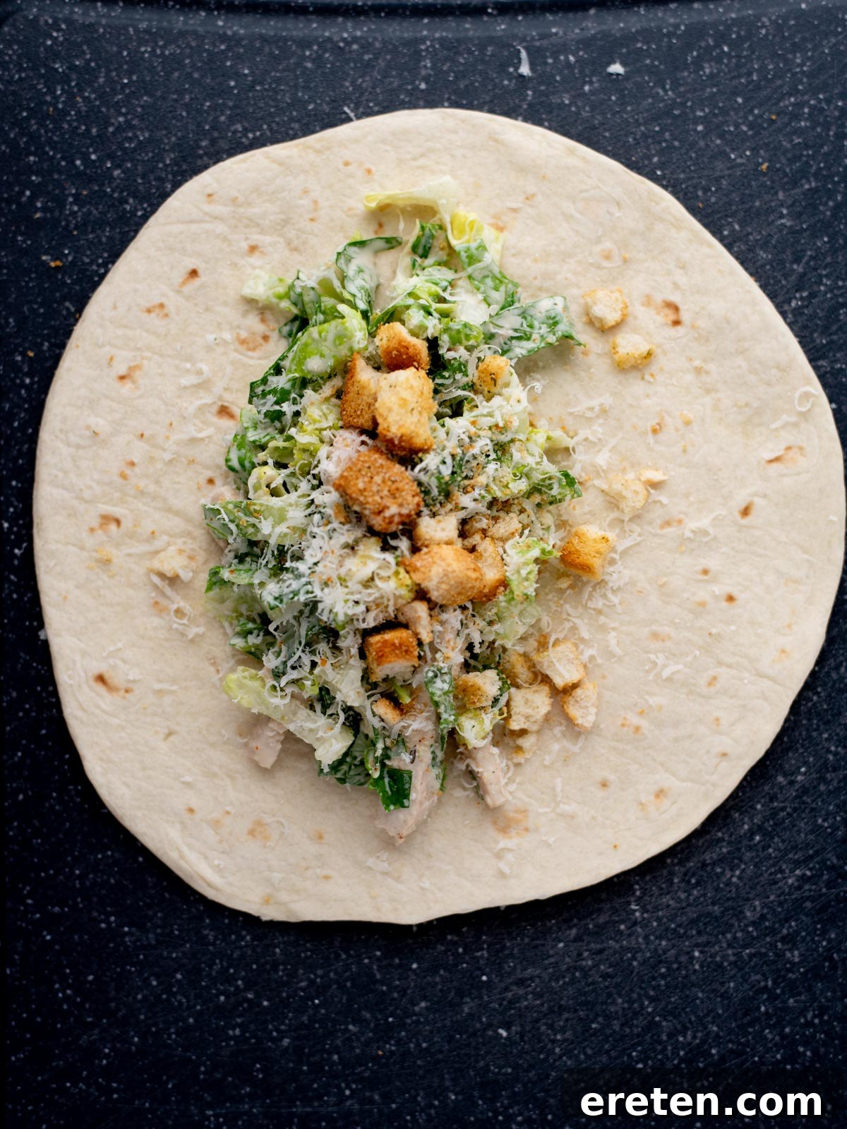 A flour tortilla on a dark surface topped with chopped romaine lettuce, shredded cheese, croutons, and pieces of chicken, ready to be wrapped as a Caesar salad wrap.