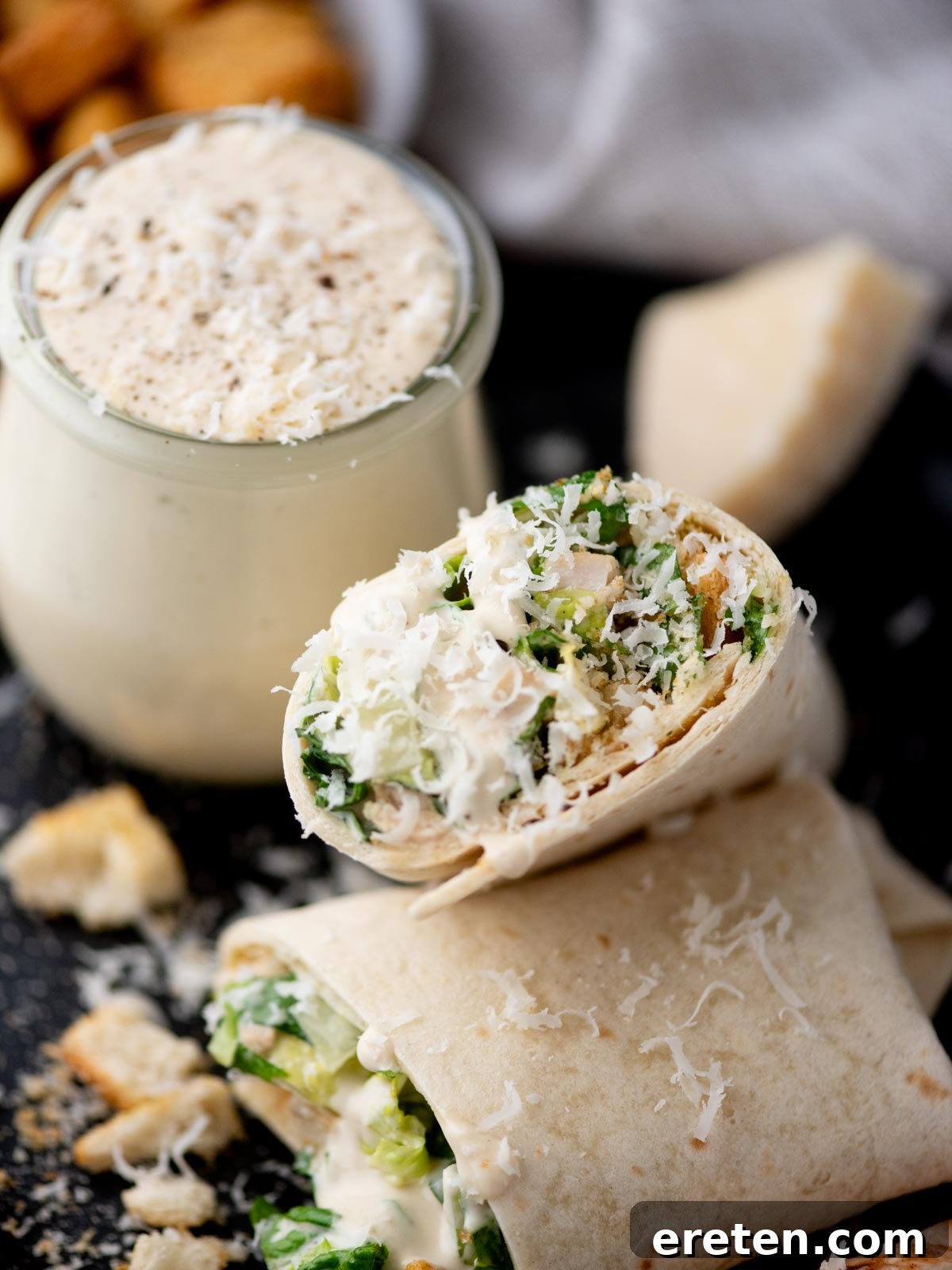 A close-up of a Caesar salad wrap cut in half, filled with lettuce, chicken, and grated cheese, with a jar of creamy Caesar dressing and croutons in the background.