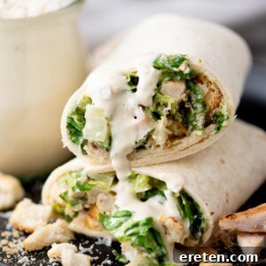 A close-up of a chicken Caesar salad wrap cut in half, filled with lettuce, chicken, croutons, and creamy dressing, with sauce drizzling out and crouton pieces scattered on the plate.