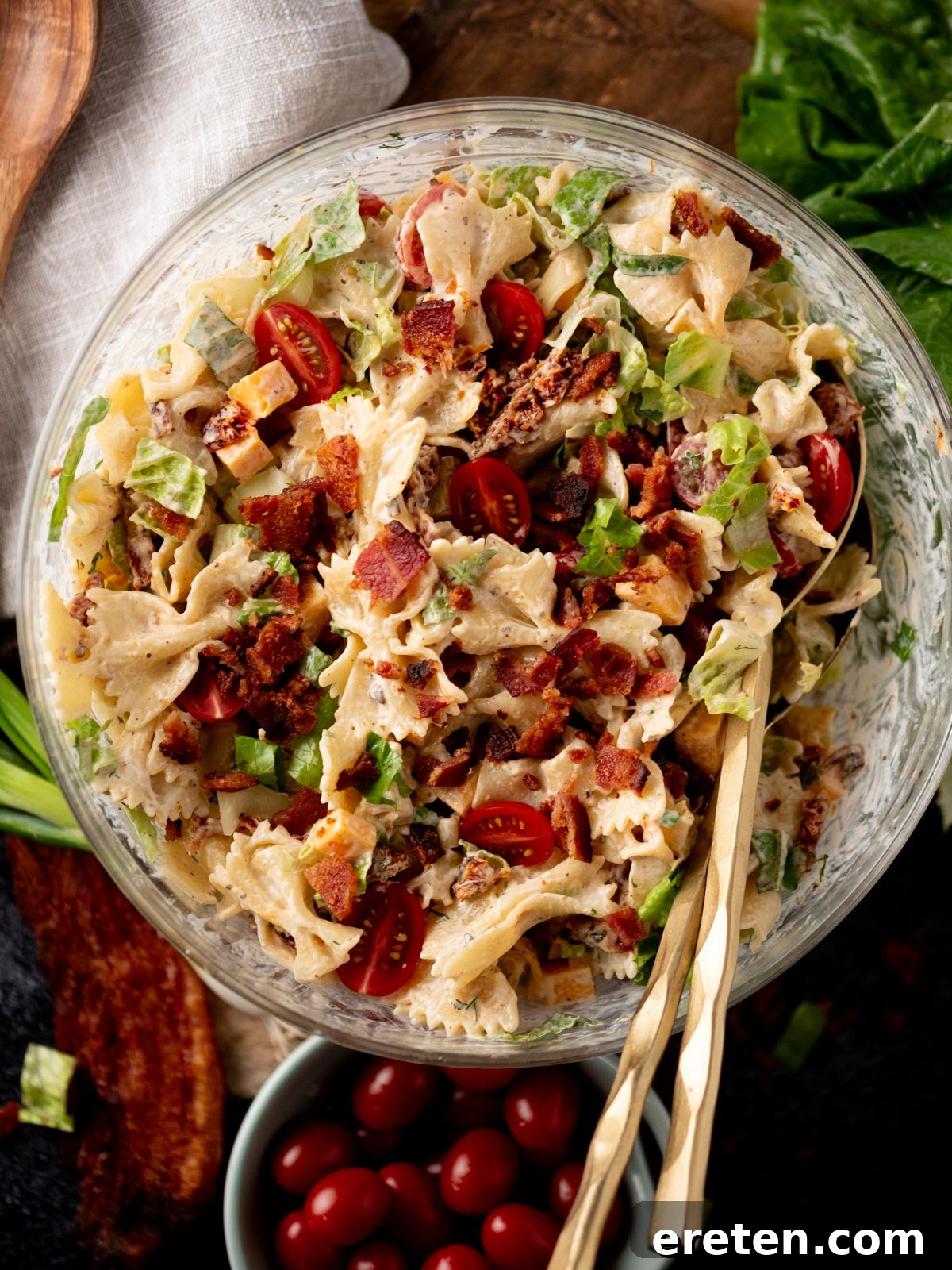 A glass bowl filled with bowtie pasta salad mixed with bacon, lettuce, cherry tomatoes, and a creamy dressing. Two gold utensils rest in the bowl, and fresh greens and tomatoes are nearby.