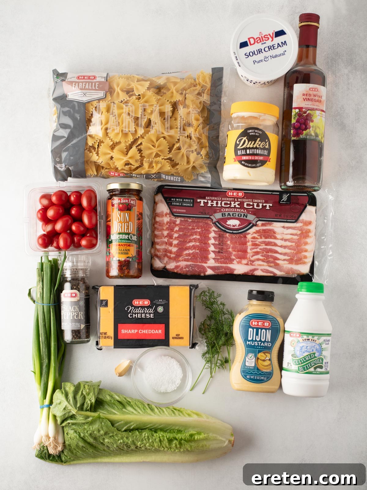 A flat lay of ingredients including bowtie pasta, sour cream, red wine vinegar, grape tomatoes, sun-dried tomatoes, thick cut bacon, sharp cheddar cheese, Dijon mustard, whole milk, green onions, fresh dill, Romaine lettuce, garlic, black pepper, and flour.