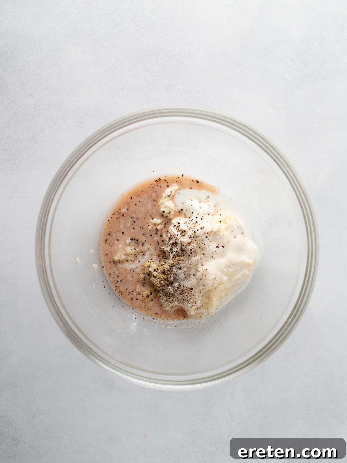 A glass mixing bowl containing mayonnaise, red liquid, and cracked black pepper on a light grey surface, viewed from above.