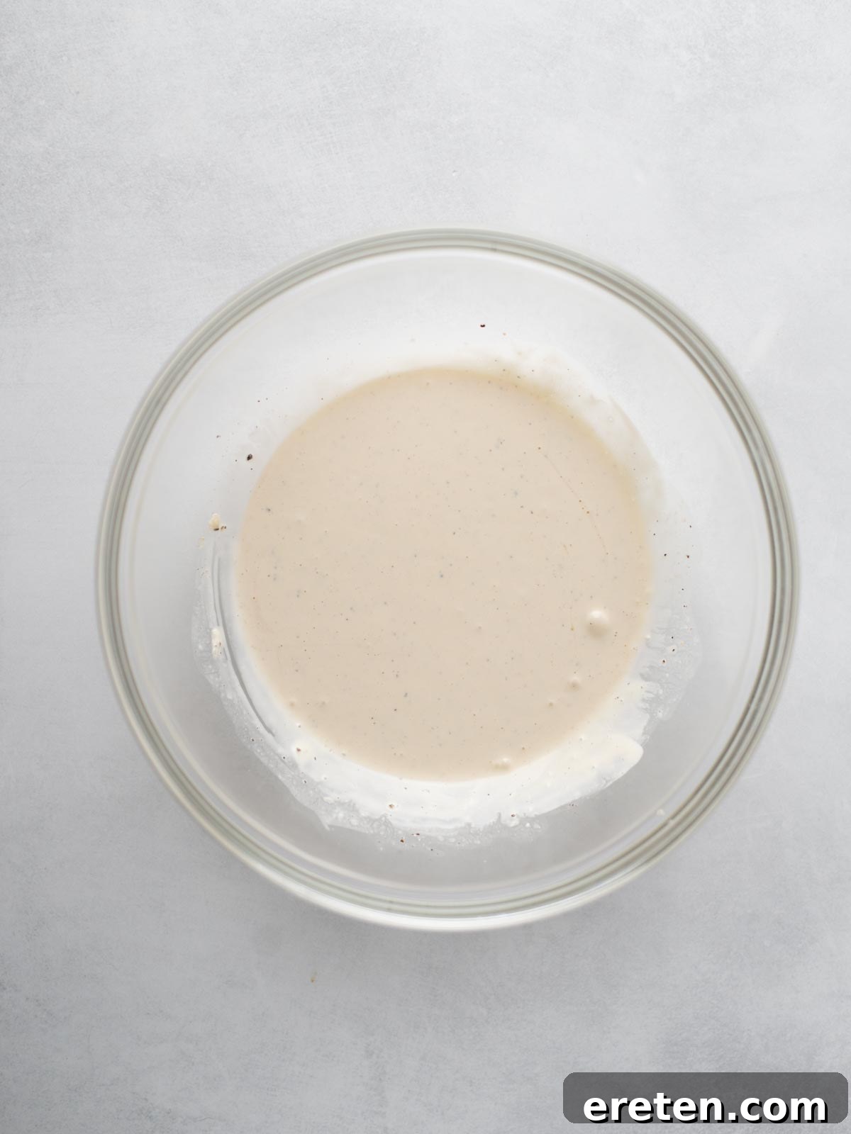 A clear glass bowl containing a small amount of light beige batter or sauce sits on a light gray surface.