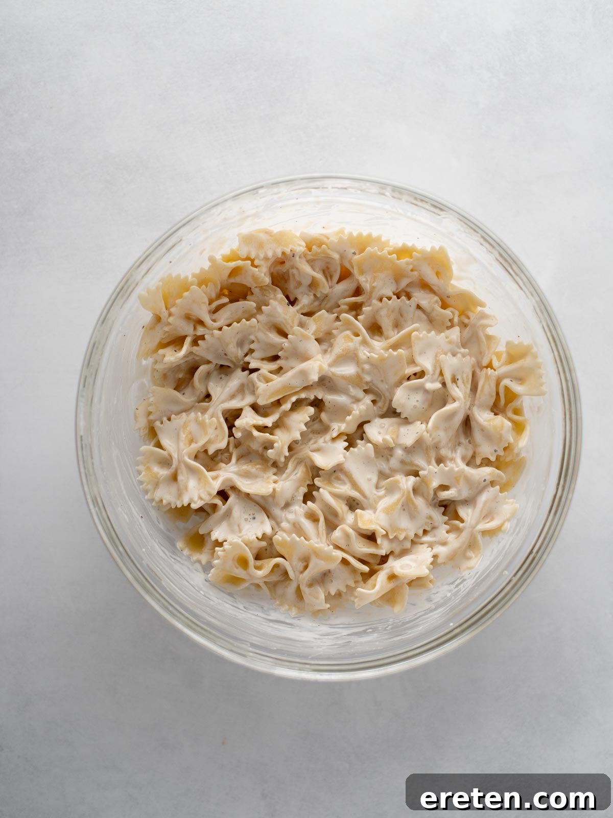 A clear glass bowl filled with farfalle pasta coated in a creamy white sauce, viewed from above on a light gray surface.