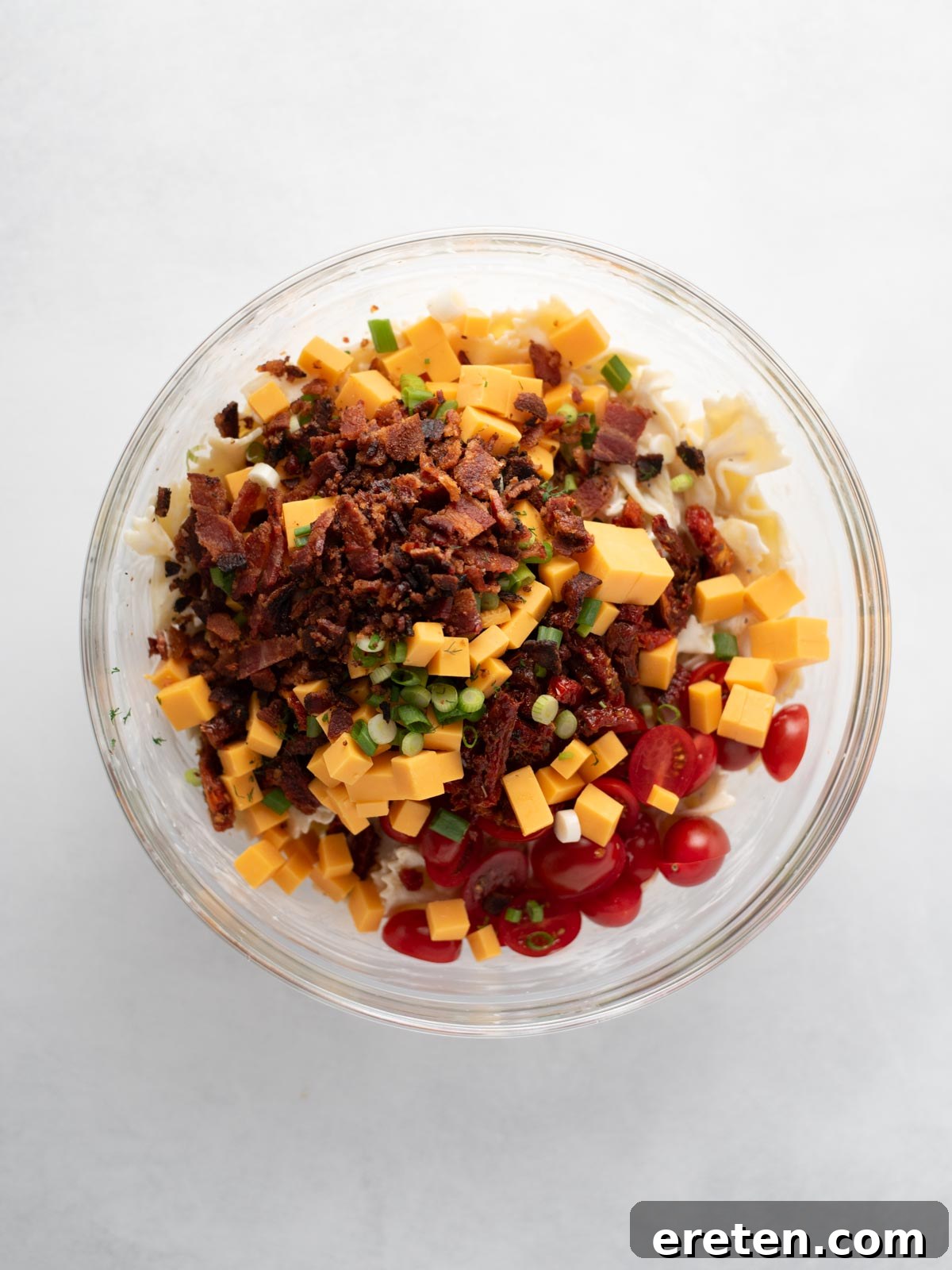 A glass bowl filled with uncooked pasta, diced cheddar cheese, bacon bits, cherry tomatoes, green onions, and herbs, photographed from above on a white background.