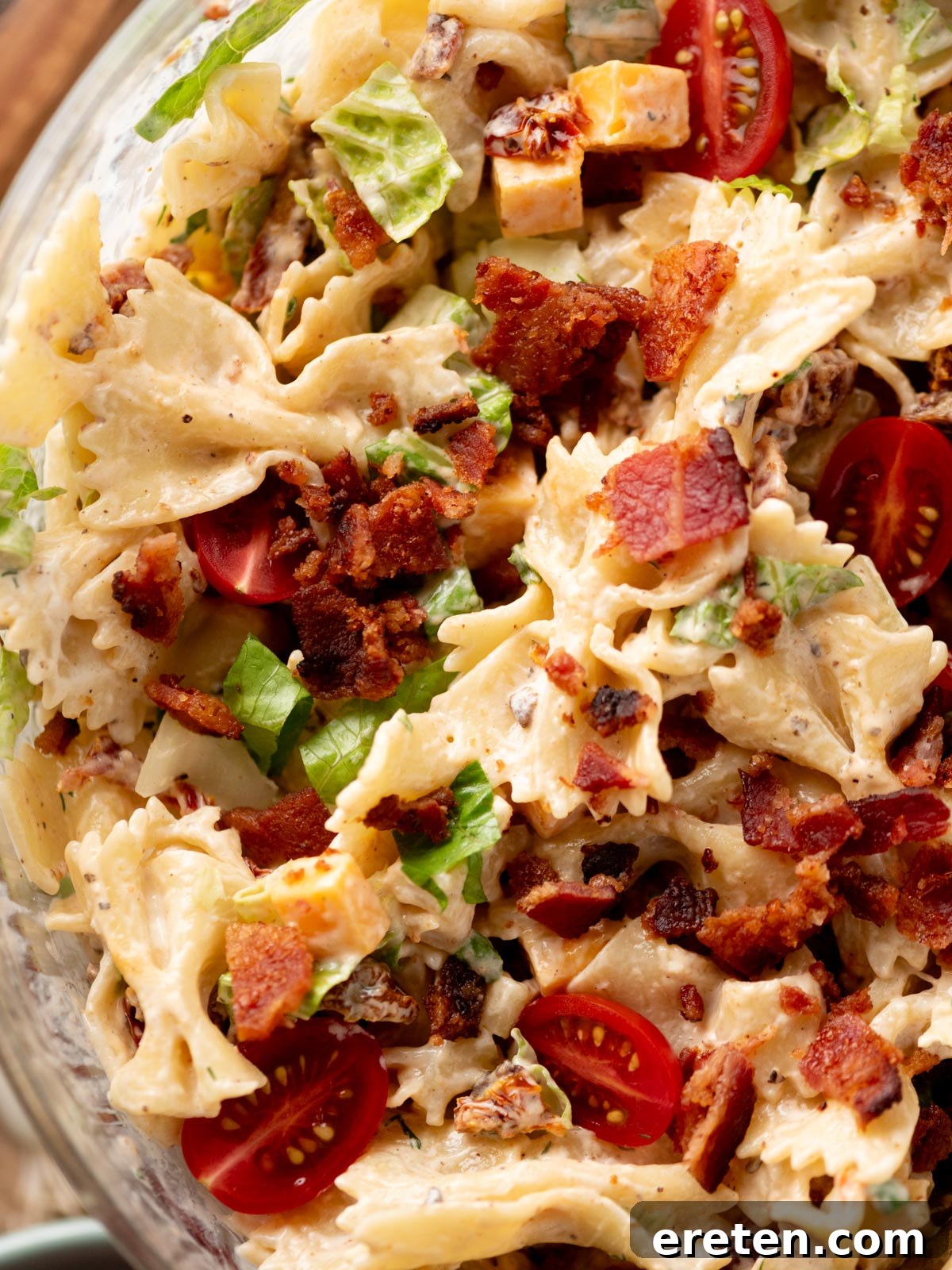 A close-up of a pasta salad with bowtie pasta, cherry tomatoes, lettuce, cubed cheese, bacon pieces, and a creamy dressing.