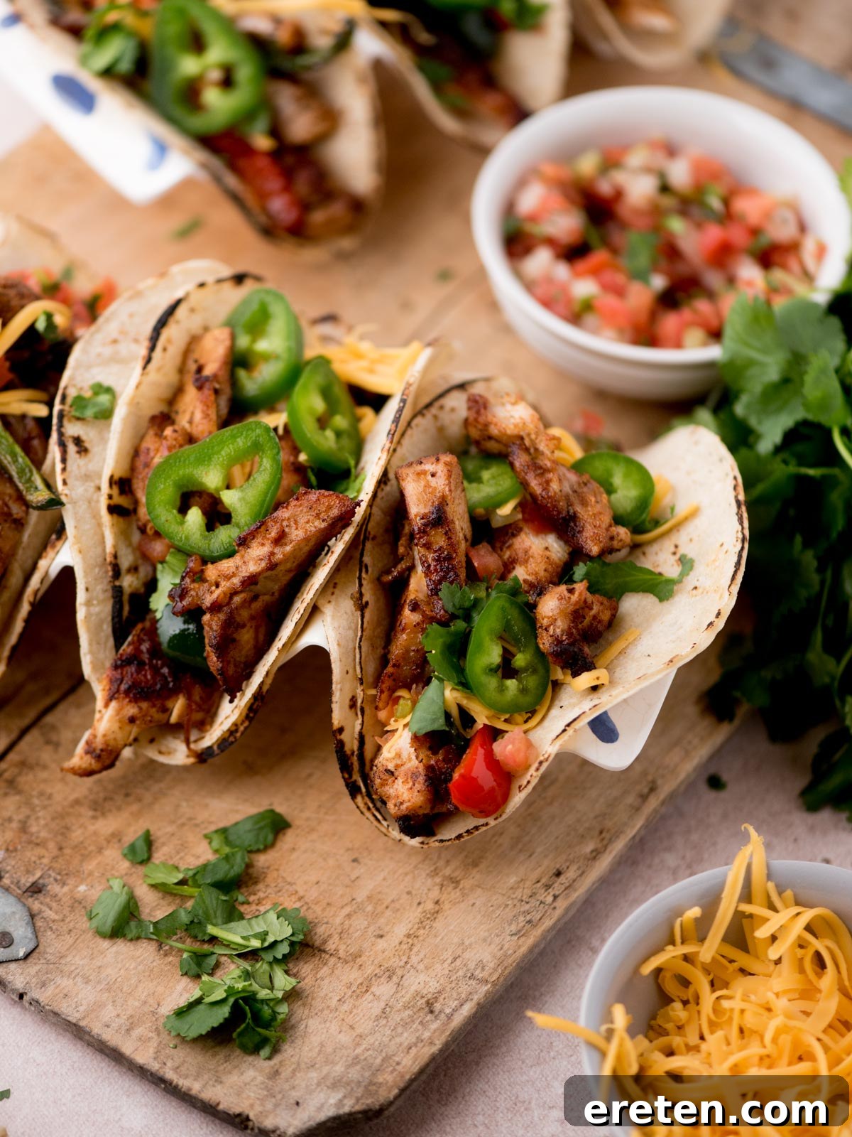 Three grilled chicken tacos, inspired by Texas chicken fajitas, topped with sliced jalapeños, shredded cheese, and diced tomatoes on a wooden board, surrounded by fresh cilantro, extra cheese, and a bowl of pico de gallo.
