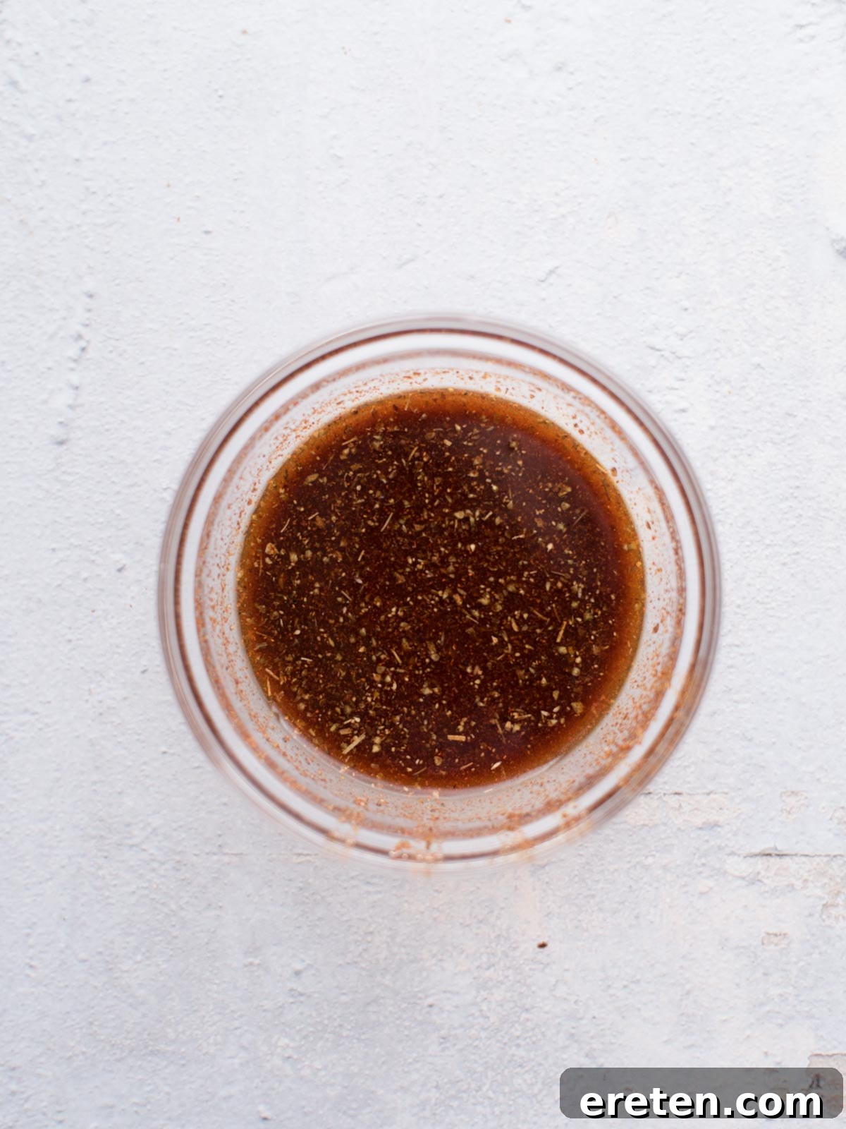 A glass bowl containing a brown, herb-speckled liquid sits on a light, textured surface. This flavorful mixture appears to be a marinade or dressing, perfect for making easy chicken fajitas.