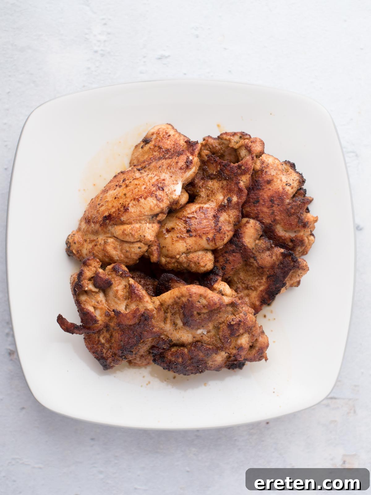 A white plate with several pieces of cooked, seasoned chicken thighs stacked on it, perfect for making easy chicken fajitas, is placed on a light-colored surface.