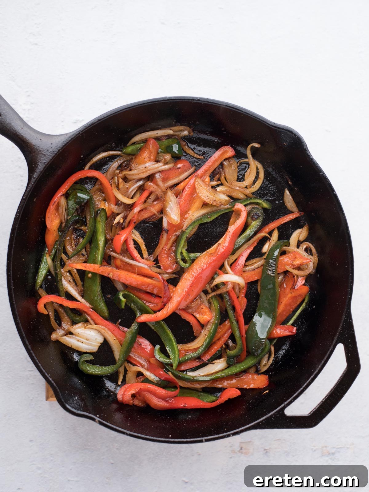 Sautéed sliced red and green bell peppers with onions in a black cast iron skillet, the perfect filling for easy chicken fajitas, viewed from above on a white background.