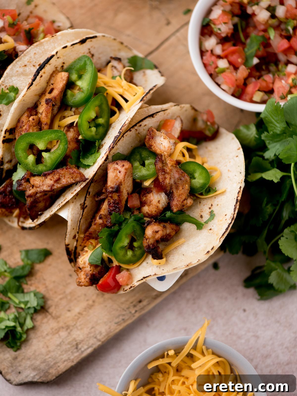 Three grilled chicken tacos inspired by Texas chicken fajitas, topped with sliced jalapeños, shredded cheese, and diced tomatoes on corn tortillas. Served on a wooden board with fresh cilantro, pico de gallo, and extra cheese.