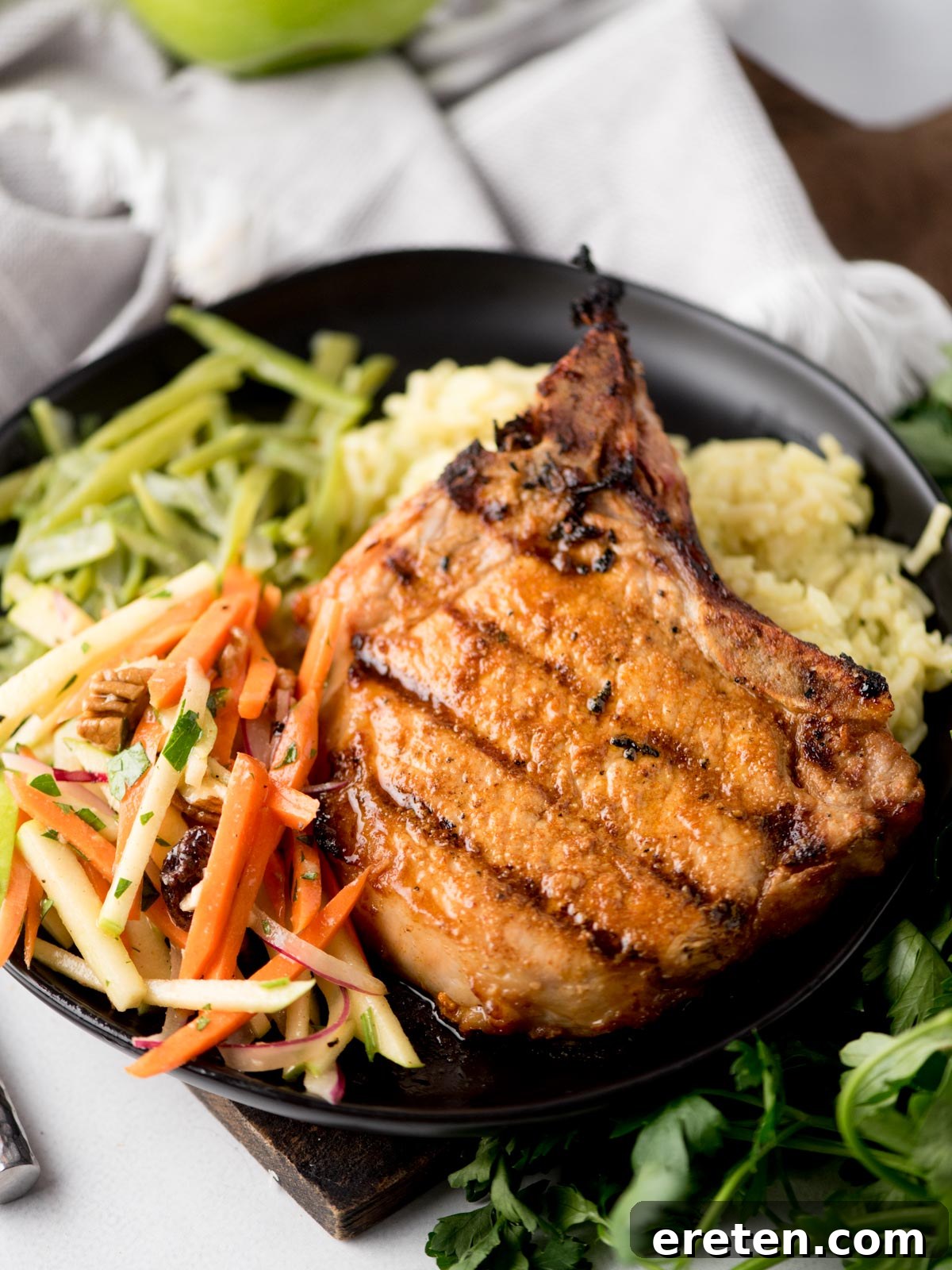 A perfectly grilled pork chop served elegantly on a black plate, accompanied by creamy mashed potatoes, vibrant green beans, and a refreshing apple slaw featuring julienned carrots and apples, all garnished with fresh herbs. This delightful presentation showcases a masterful way to enjoy grilled pork chops.