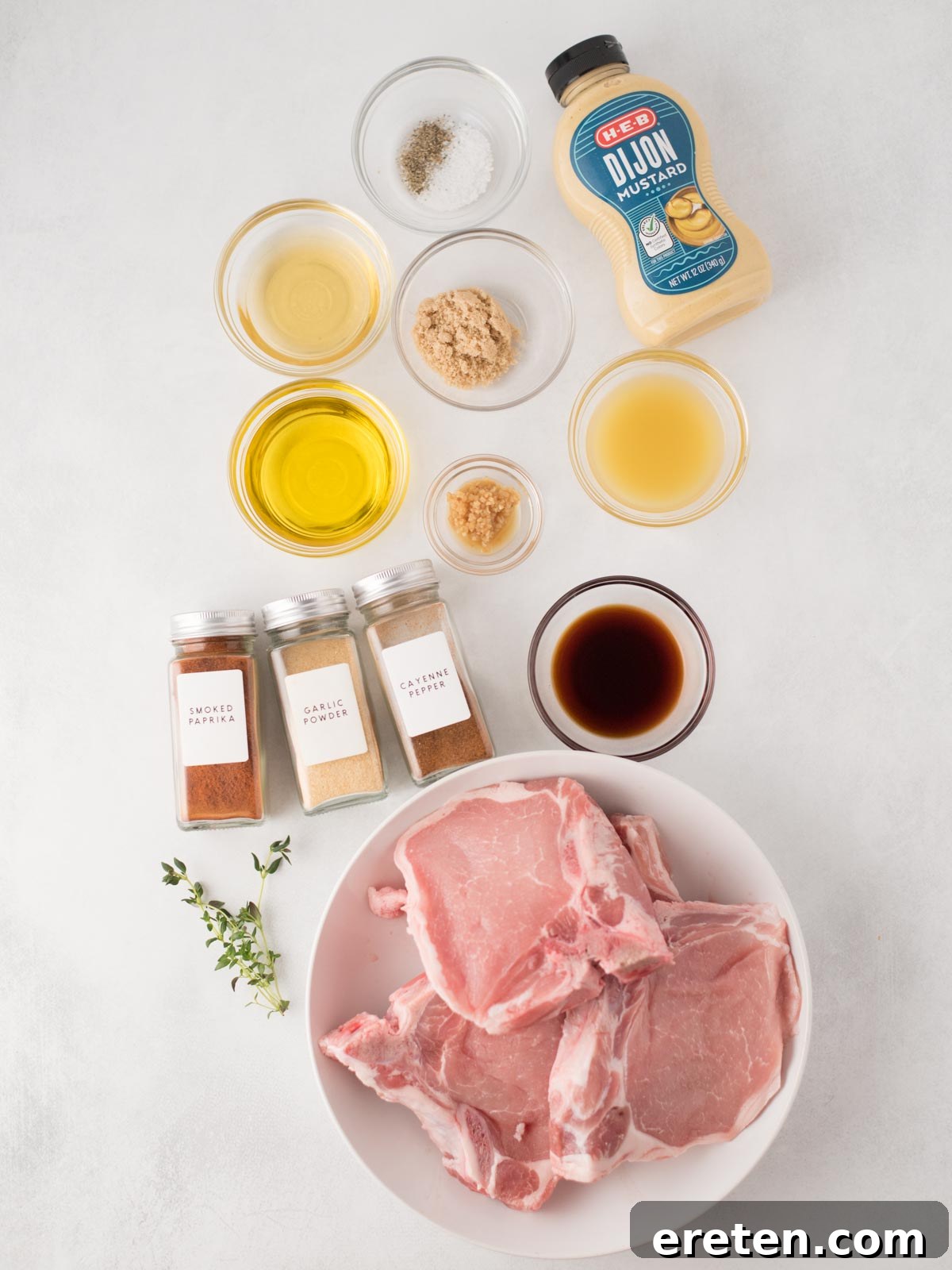 A bowl of raw pork chops, perfectly prepared for grilling, is surrounded by an array of small bowls containing fresh ingredients: olive oil, minced garlic, Dijon mustard, soy sauce, apple cider vinegar, salt, pepper, and three distinct spice jars. All are neatly arranged on a pristine white surface, highlighting the fresh components for this grilled pork chops recipe.