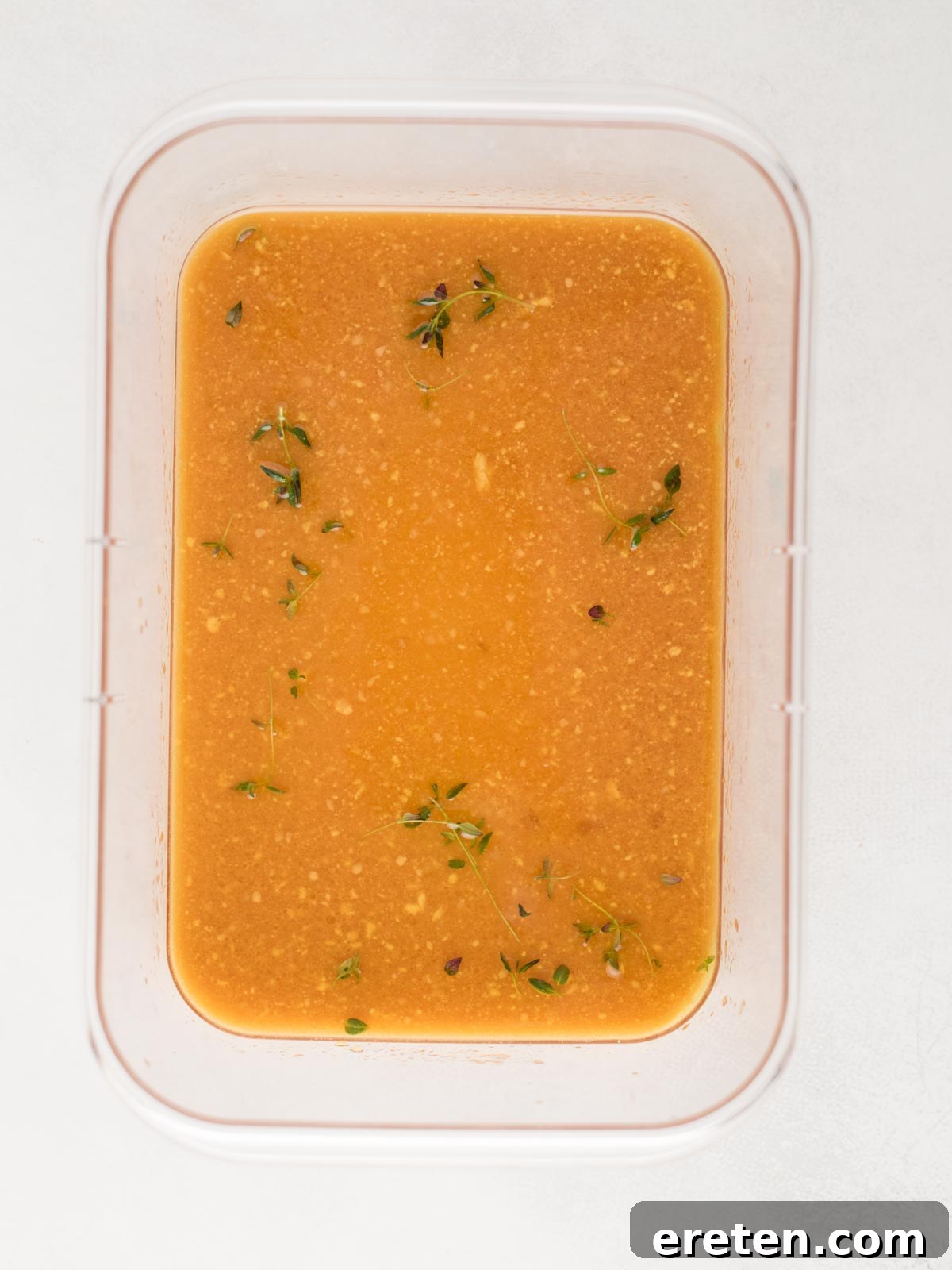 A rectangular container filled with a rich orange-brown marinade, speckled with small pieces of fresh herbs and spices, specifically prepared for grilled pork chops. A few sprigs of fresh thyme float on the surface, adding to the aromatic appeal. The container is set against a clean white background, highlighting its contents.