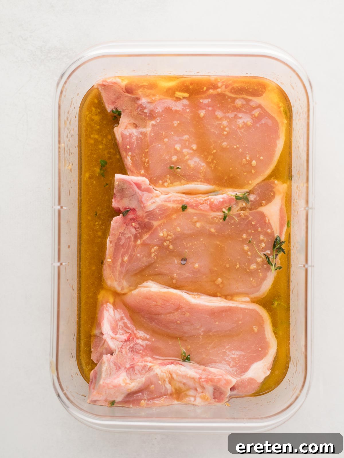 Three raw pork chops, ready to be transformed into succulent grilled pork chops, are perfectly immersed in a light brown marinade within a rectangular container. The marinade is dotted with small bits of herbs and garlic, infusing flavor into the meat. The container rests on a clean white surface, emphasizing the marinating process.