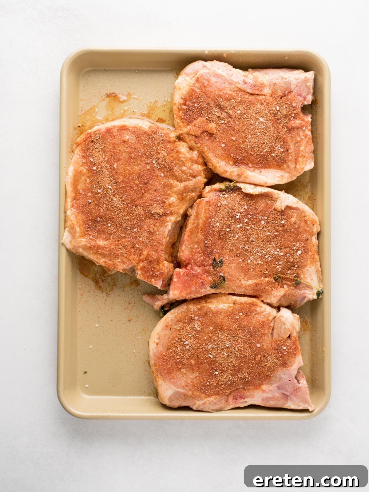 A baking tray holds four raw pork chops, each liberally sprinkled with seasoning, arranged neatly on a light surface. This image perfectly captures the preparation stage for creating delicious grilled pork chops.
