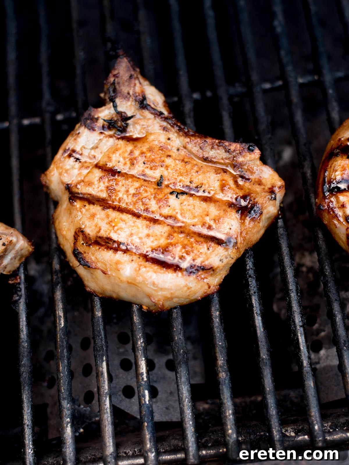 A succulent grilled pork chop, featuring prominent dark grill marks, cooking on a sturdy metal barbecue grill. The charred lines and golden-brown exterior make for an irresistible sight, showcasing the artistry of perfectly grilled pork chops.