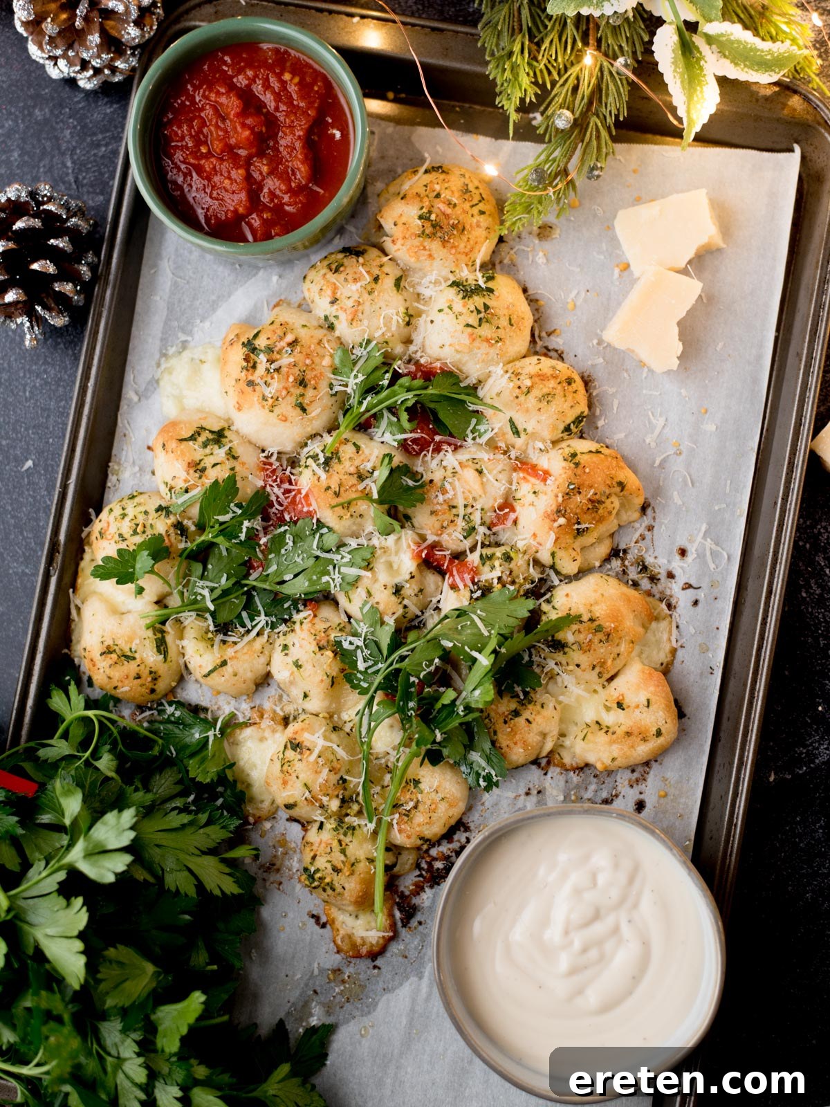 A festive, tree-shaped pull-apart bread with herbs and cheese on a baking tray, served with tomato sauce and a creamy dip, garnished with fresh parsley and Parmesan cheese. Holiday decor surrounds the tray.