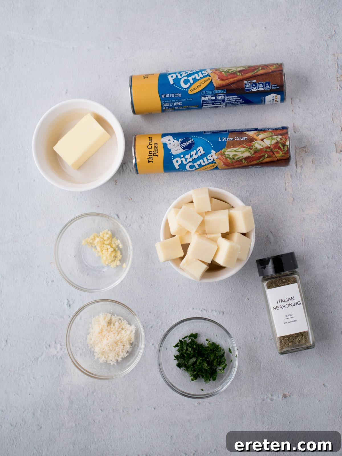 Ingredients on a light surface: two cans of pizza crust, a bowl of butter, cubed cheese, grated cheese, minced garlic, chopped herbs, and a jar labeled Italian Seasoning.