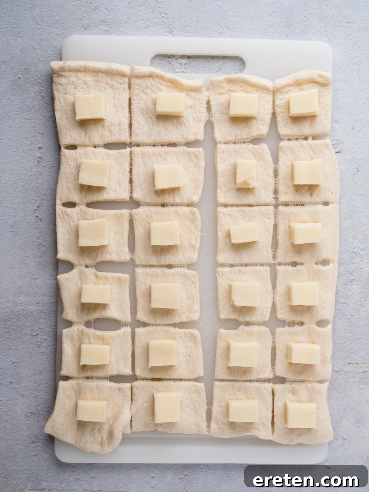 A white cutting board with rectangular dough pieces, each topped with a small square of cheese, arranged in a grid pattern on a light gray surface.