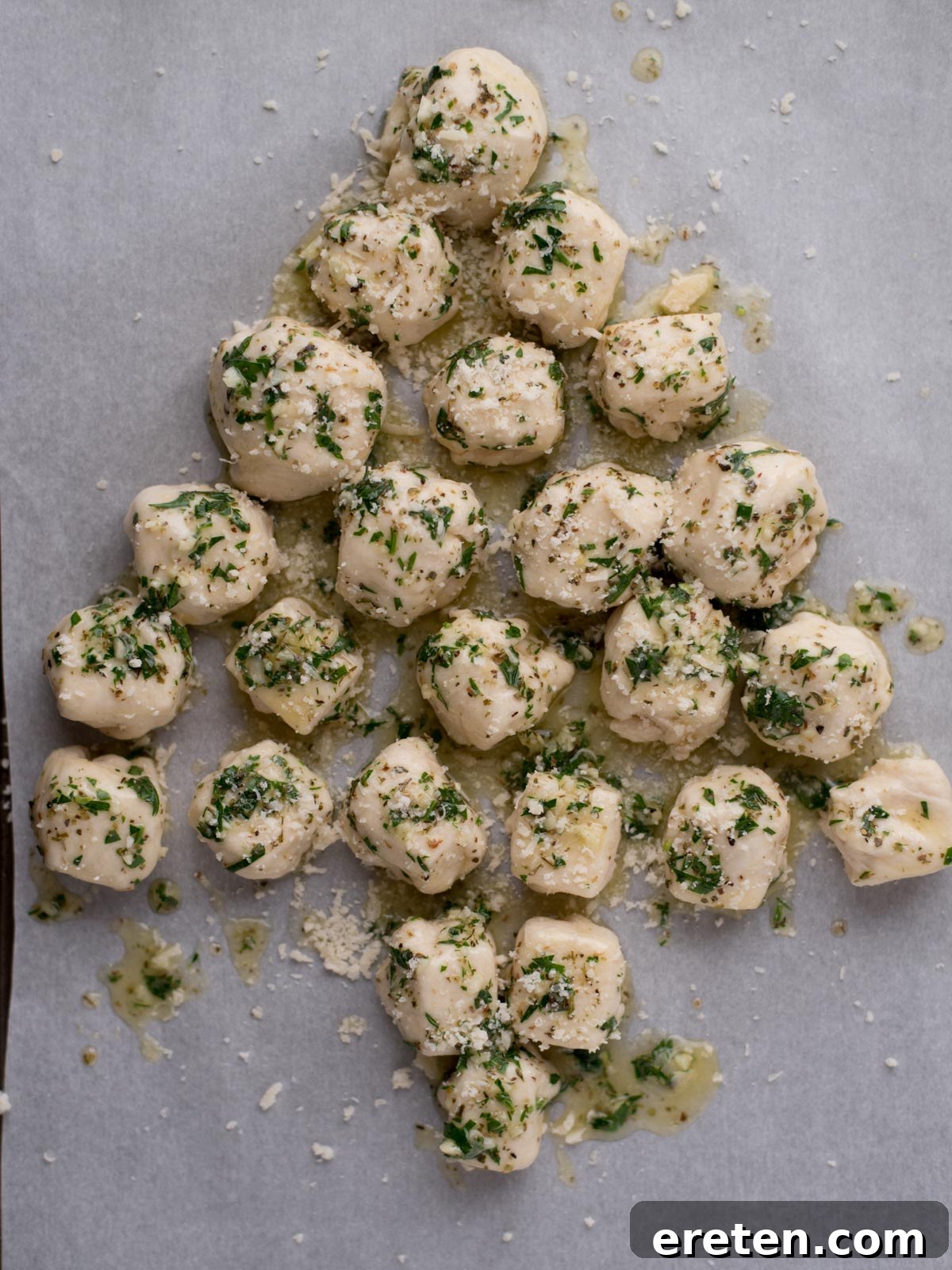 Balls of dough topped with chopped herbs and grated cheese are arranged on a baking sheet in the shape of a Christmas tree.
