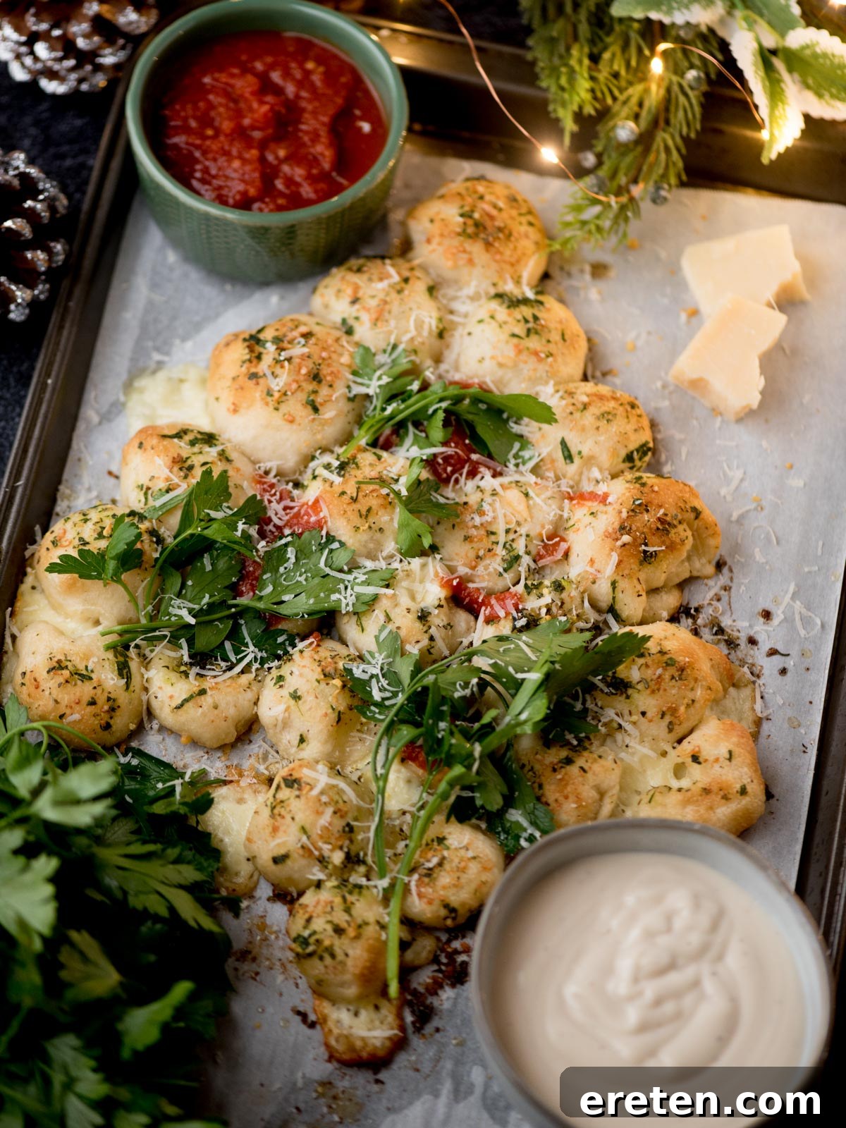 A tray of pull-apart garlic bread bites topped with herbs and grated cheese, garnished with fresh arugula, served with bowls of marinara sauce and creamy dip, and pieces of parmesan on the side.