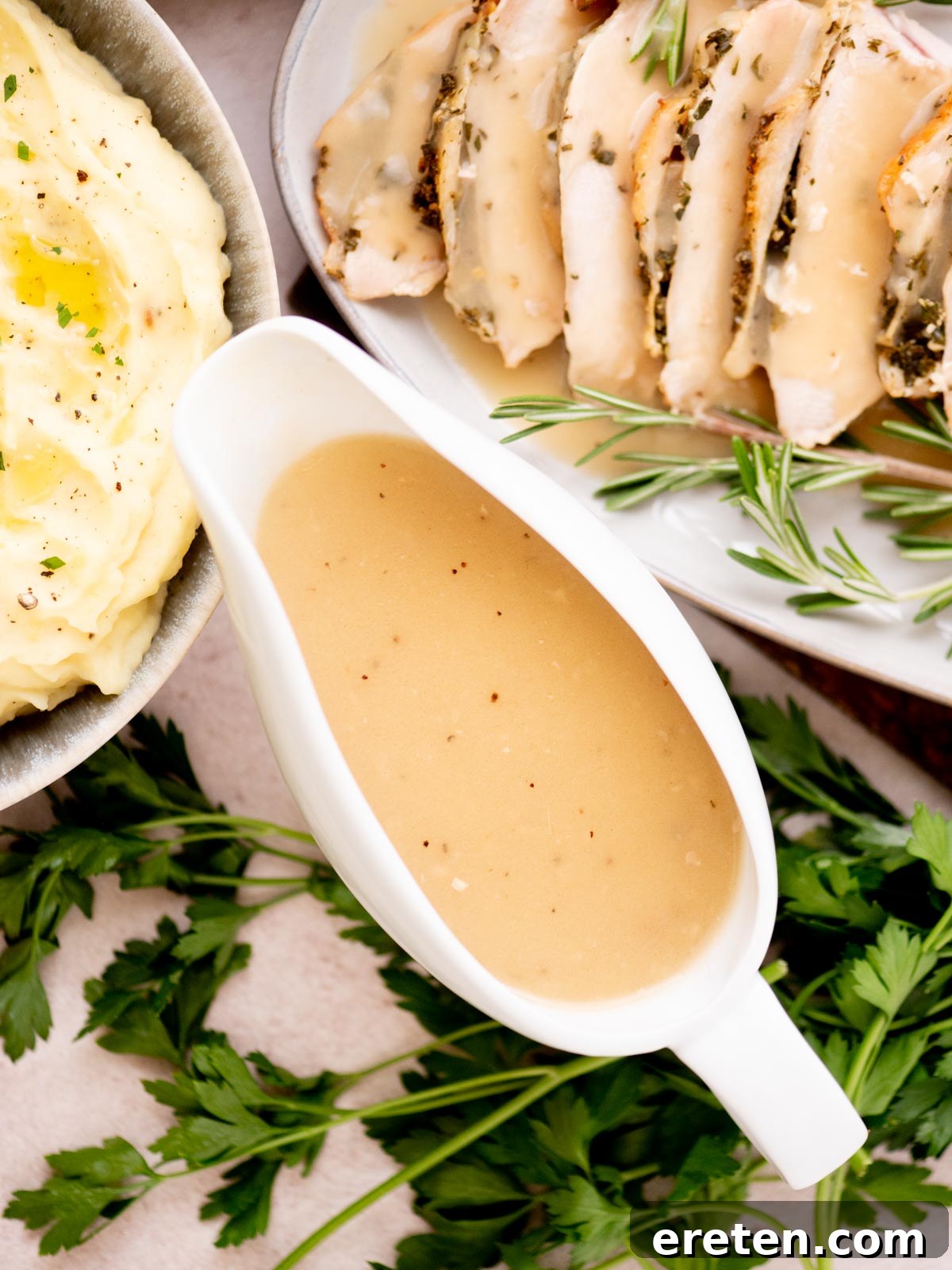 A white gravy boat filled with creamy brown Thanksgiving gravy sits on a table next to a bowl of mashed potatoes and a plate of sliced roast chicken garnished with fresh rosemary and parsley.