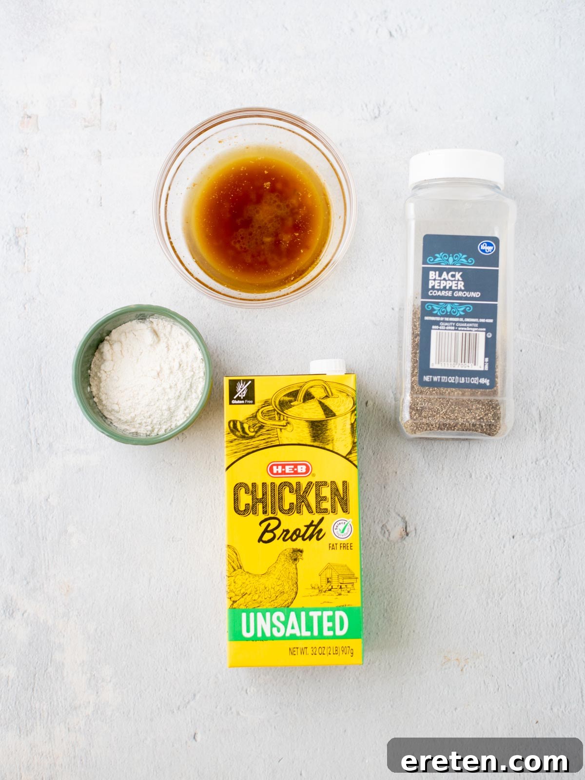 A bowl of brown liquid turkey gravy with drippings, a small bowl of white flour, a carton of unsalted chicken broth, and a container of ground black pepper are arranged on a light surface.