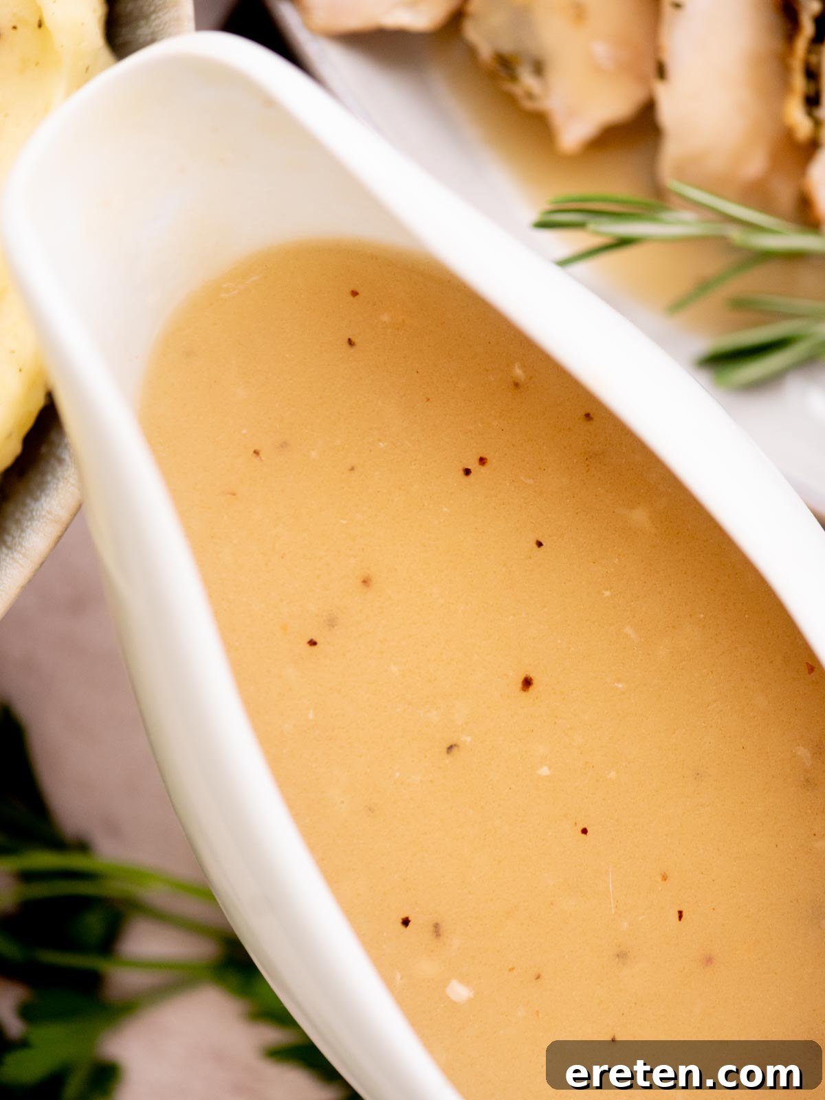 A close-up of a white gravy boat filled with creamy brown turkey gravy with drippings, speckled with pepper. Fresh herbs and a plate of sliced meat are slightly blurred in the background.