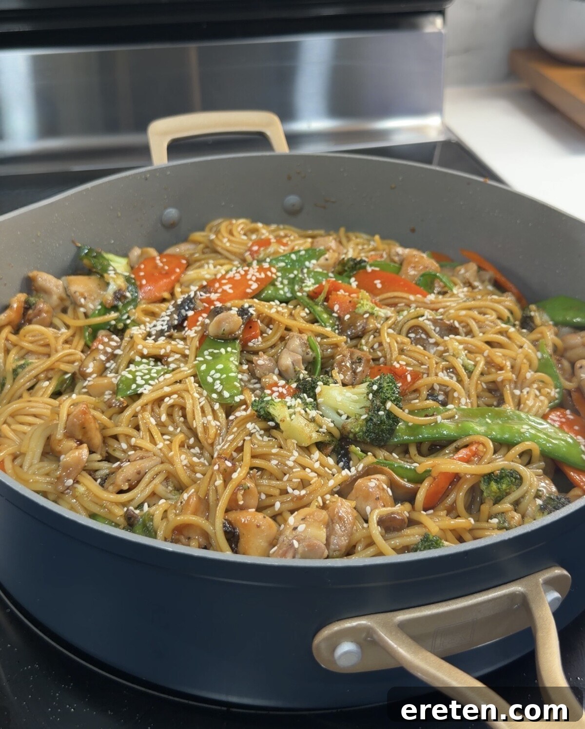 A large skillet brimming with homemade Chicken Teriyaki Noodles, featuring succulent chicken pieces, crisp broccoli florets, vibrant snap peas, sweet carrots, and earthy mushrooms, all generously garnished with toasted sesame seeds and ready to serve on a kitchen stove.