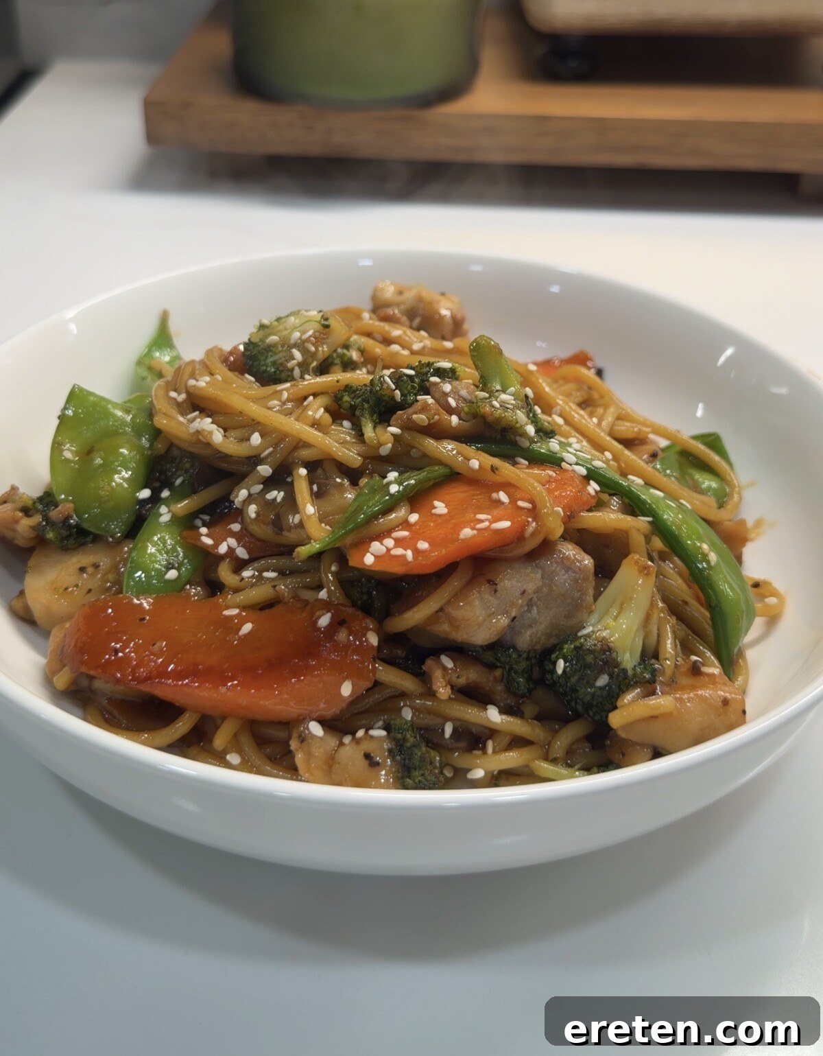 A perfectly portioned serving of Homemade Chicken Teriyaki Noodles in a pristine white bowl, showcasing succulent chicken pieces, vibrant broccoli, crisp carrots, and fresh snap peas, all generously garnished with white sesame seeds, set against a clean white countertop, ready for enjoyment.