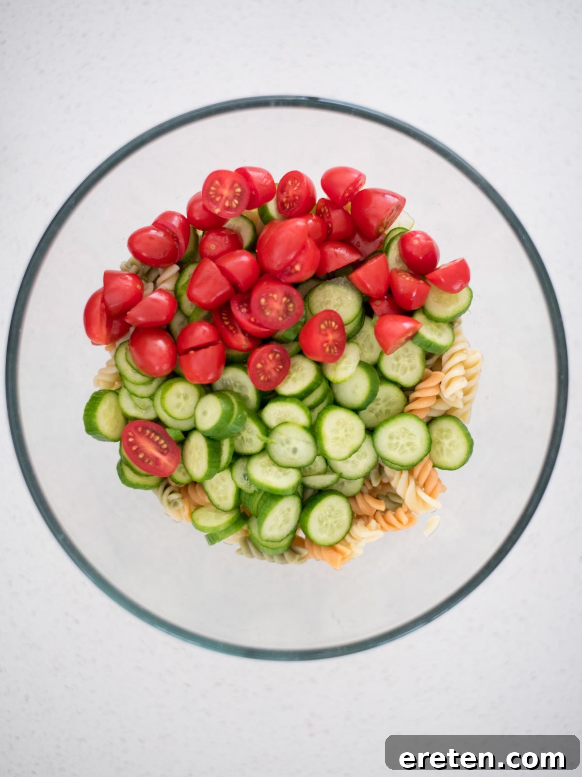 Cooked rotini pasta mixed with colorful chopped vegetables like tomatoes and cucumbers in a large bowl
