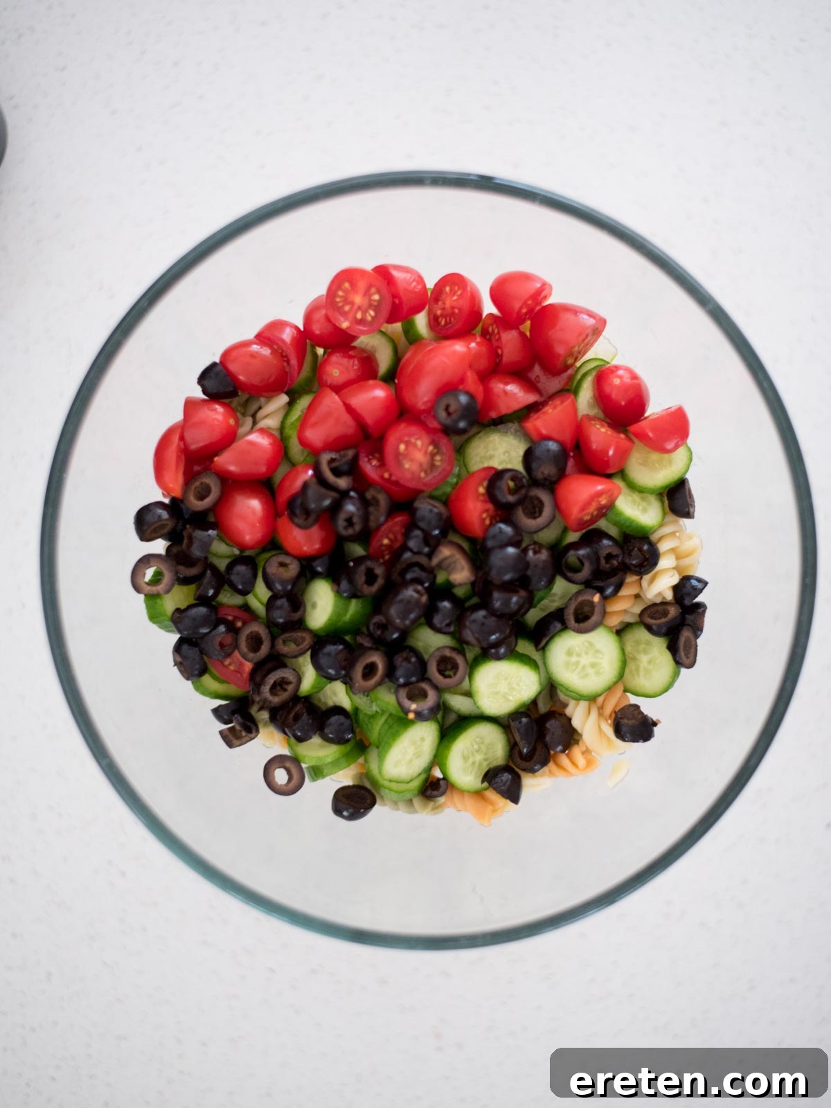 Sliced black olives being added to a large mixing bowl with cooked pasta and fresh vegetables for a Zesty Italian Pasta Salad