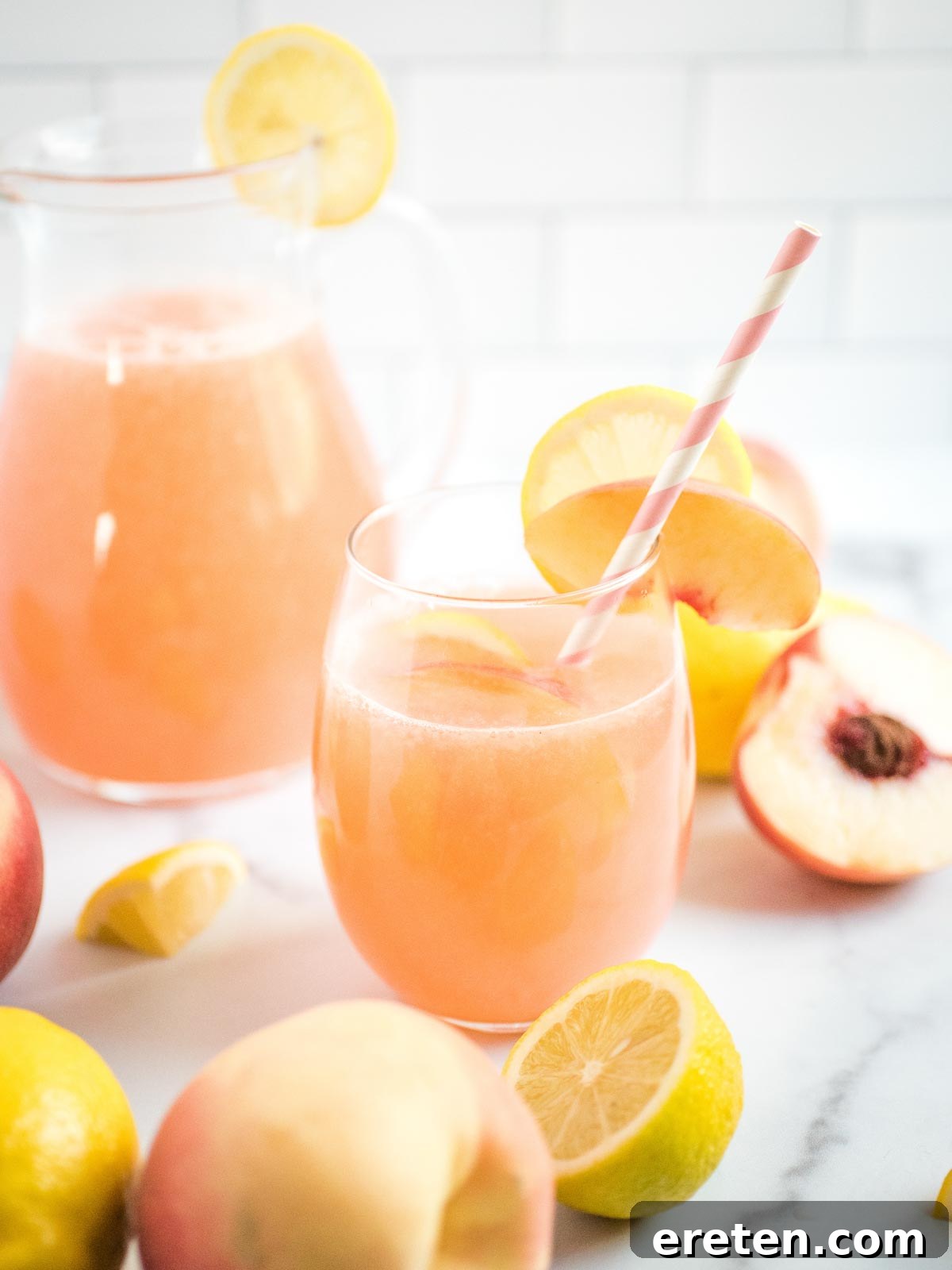A refreshing glass of peach lemonade in the foreground, with a large pitcher filled with more lemonade, fresh peaches, and lemons in the background, all on a kitchen counter.