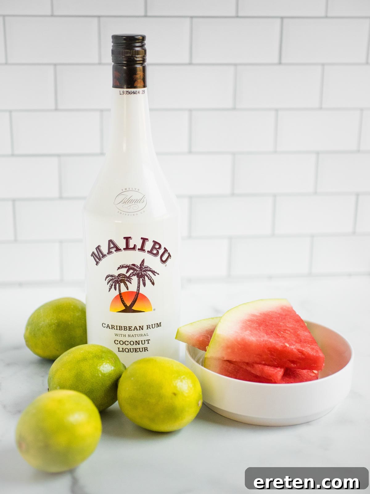 Ingredients for boozy watermelon slush laid out on a table, including a sliced watermelon, limes, a bag of sugar, and a bottle of Malibu rum.