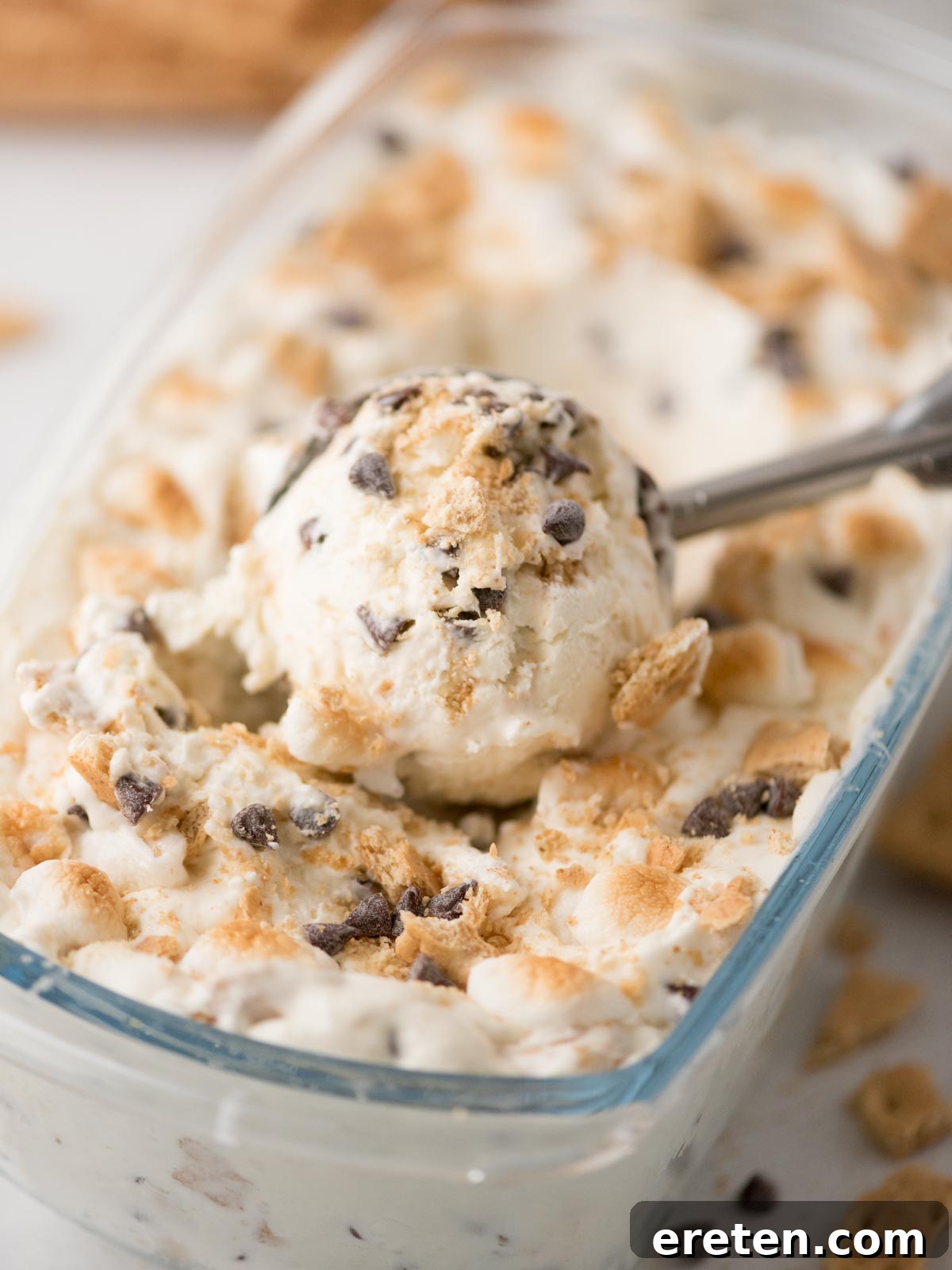 Easy Homemade Smores Ice Cream 2 Close-up of a glass dish filled with creamy ice cream mixed with chocolate chips and crushed graham crackers, with a metal scoop lifting a portion.