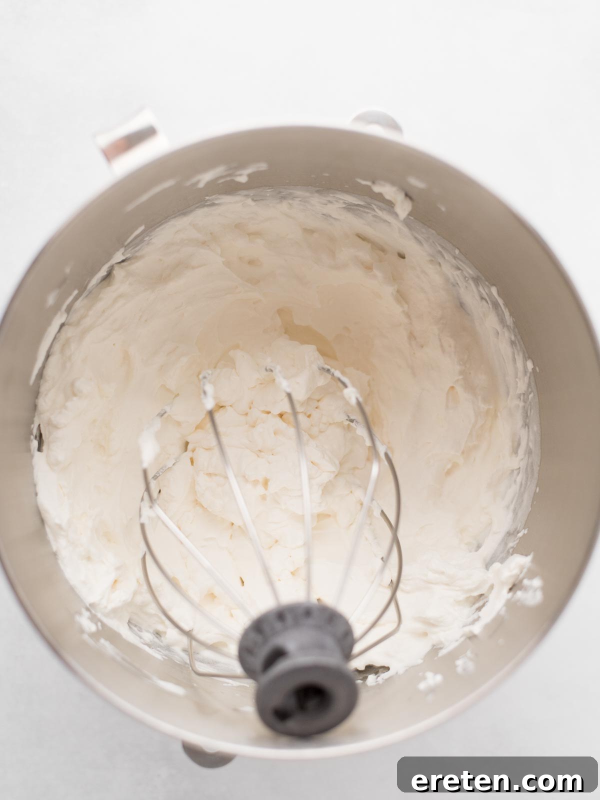 Easy Homemade Smores Ice Cream 4 A metal mixing bowl with a whisk attachment contains freshly whipped cream, with soft peaks visible and some cream clinging to the sides of the bowl. The background is a light, neutral color.