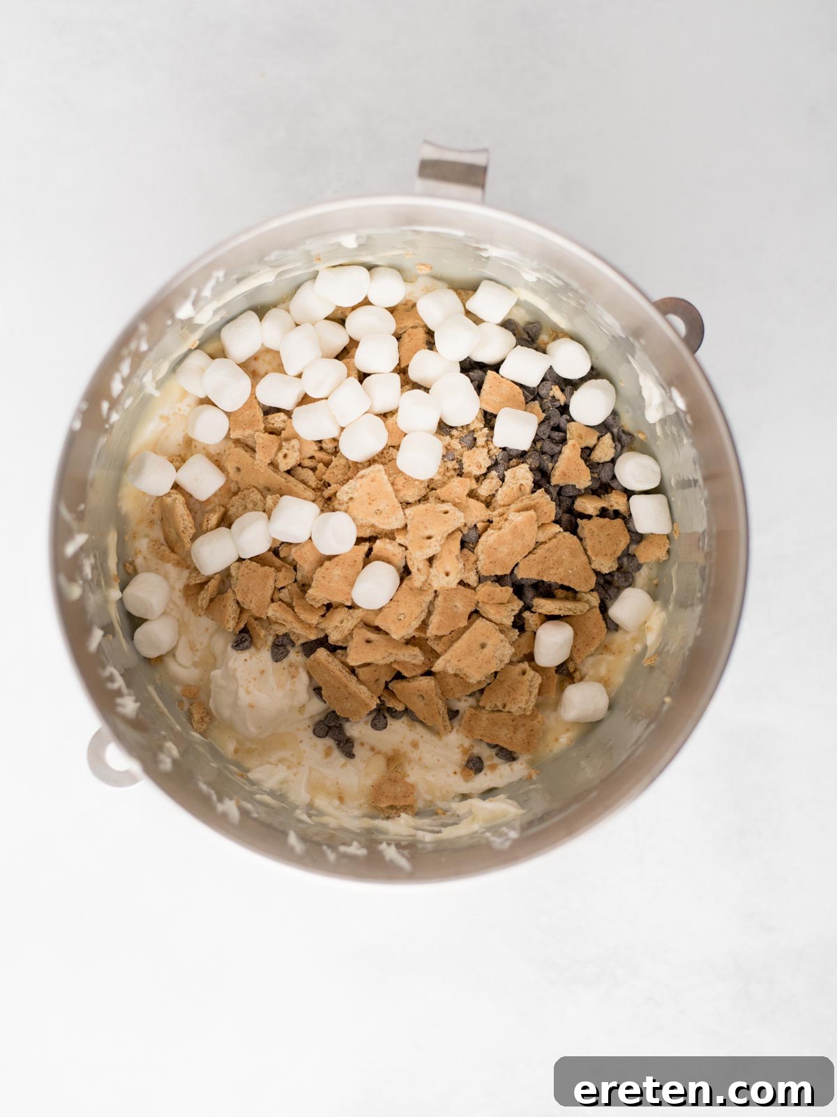 Easy Homemade Smores Ice Cream 6 A mixing bowl containing mini marshmallows, chocolate chips, and broken graham crackers on top of a creamy mixture, viewed from above.