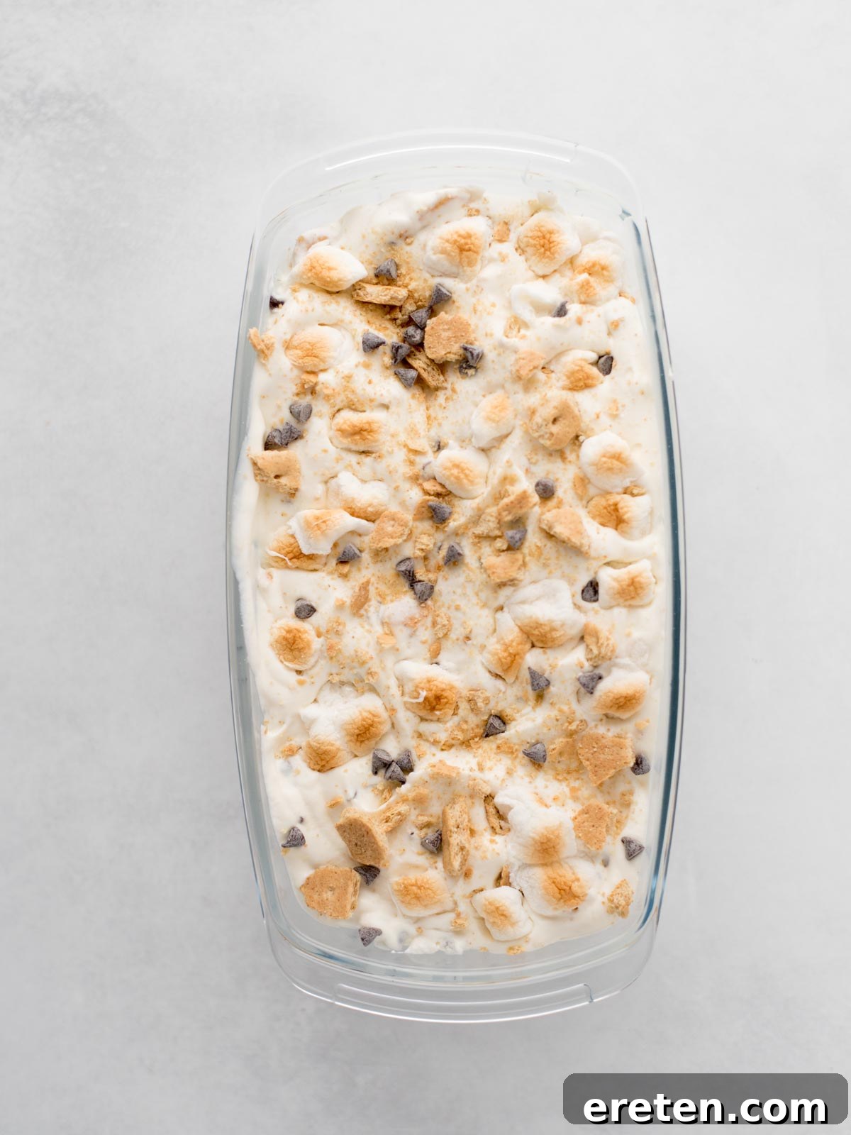 Easy Homemade Smores Ice Cream 7 A glass dish filled with ice cream topped with mini marshmallows, chocolate chips, and crushed graham crackers, resembling a smores dessert, on a light gray surface.